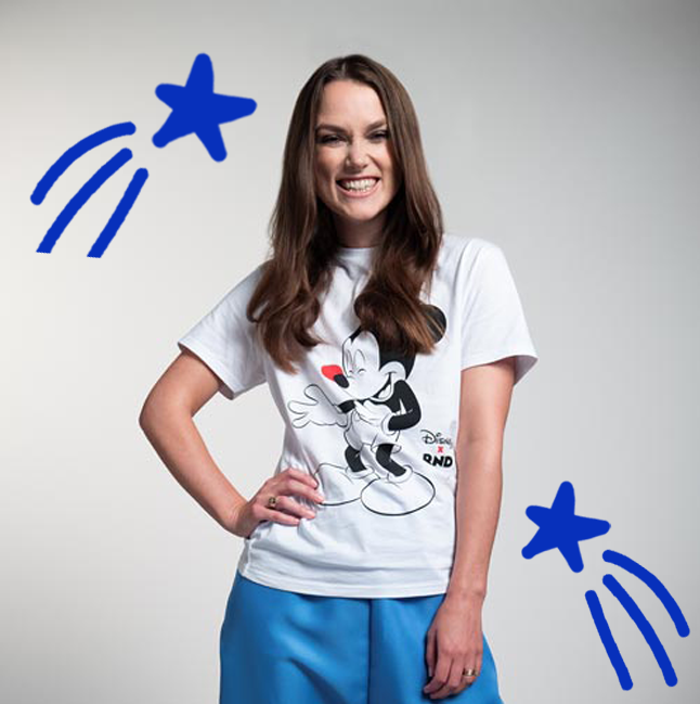 Young woman smiling in a Mickey Mouse T-shirt and blue pants, standing against a plain background with two blue cartoon-style stars and lines near her.