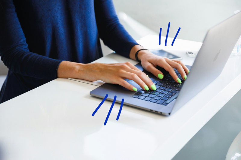 Person typing on a laptop with neon green nails, sitting at a white desk.