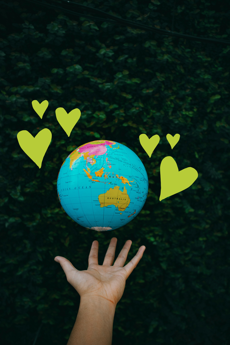 A hand holding a globe of Earth in front of a dark green leafy background, with green heart illustrations floating around the globe.