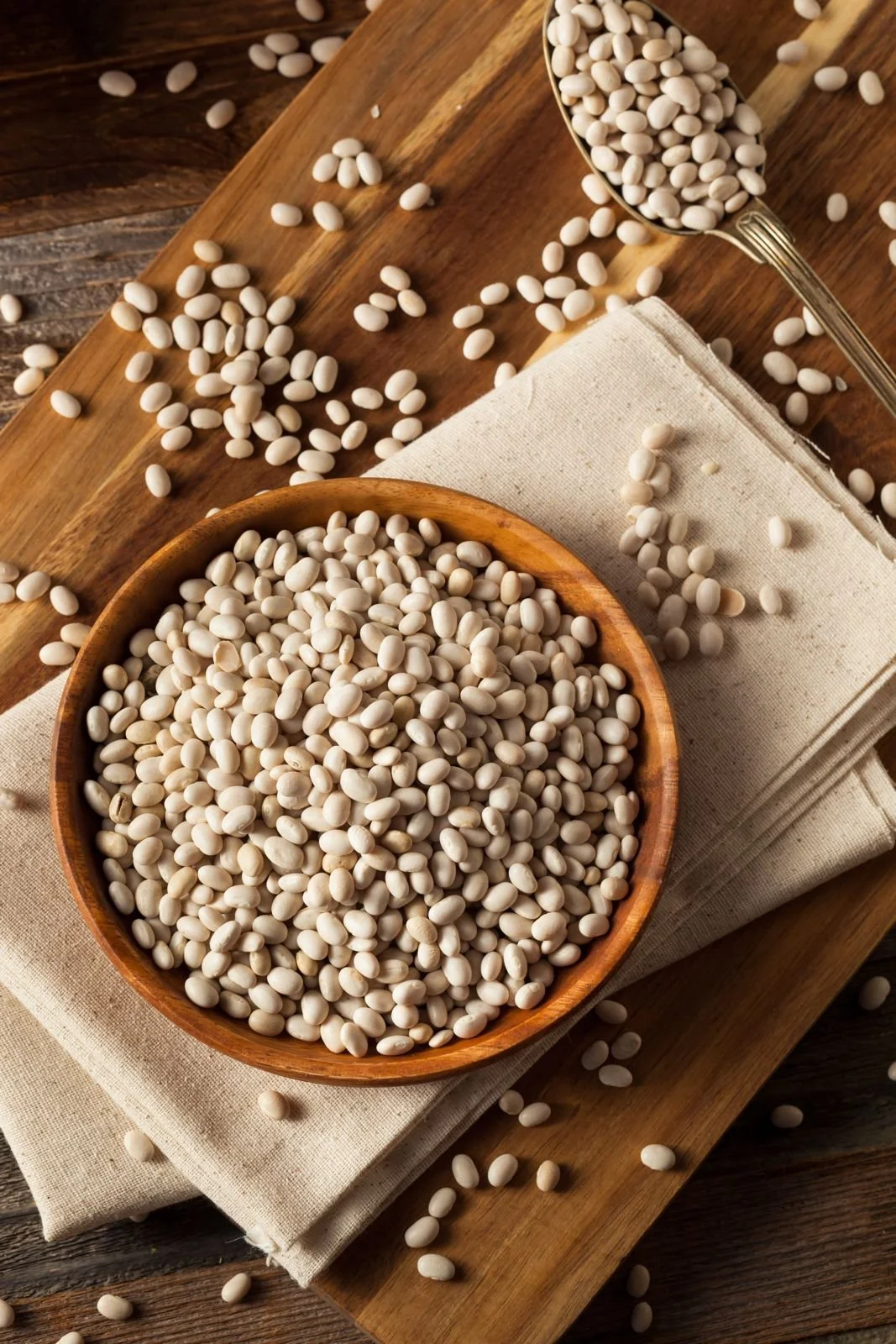 A wooden bowl filled with white beans on a wooden surface, with some beans spilled around the bowl, a metal scoop also filled with beans, and a white cloth underneath the bowl.