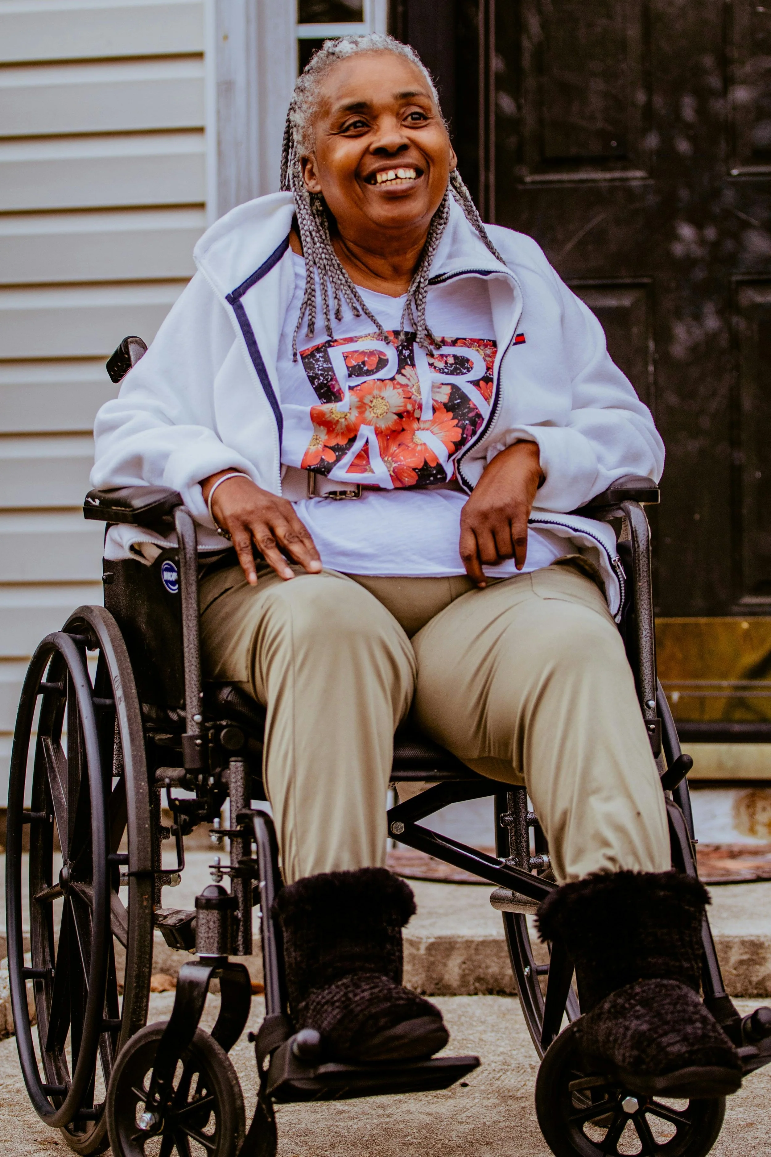 An older woman with gray hair and a bright smile sitting in a wheelchair outside a house, wearing a white jacket over a colorful shirt with floral patterns.