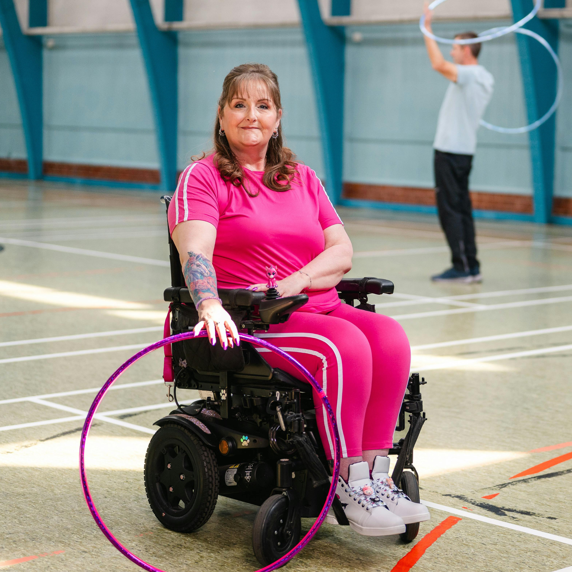 A woman in a pink outfit with floral tattooed arm sitting in a wheelchair holding a hula hoop, with a young man in a white shirt in the background also playing with a hula hoop inside a gymnasium.