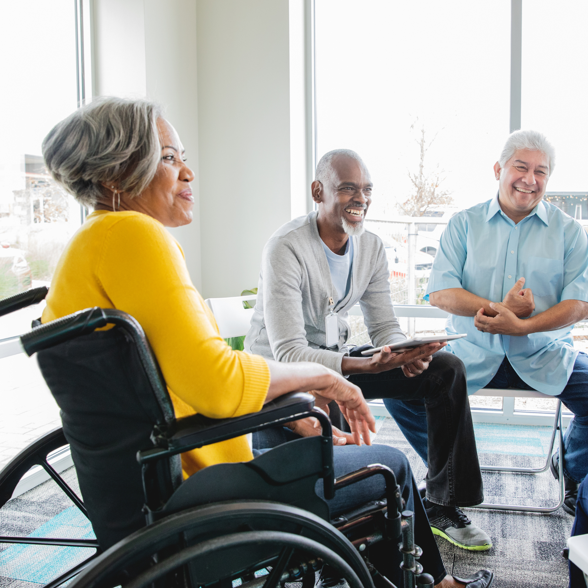 A diverse group of four people, three men and one woman, sitting and standing in a bright room with large windows, engaging in conversation and smiling.