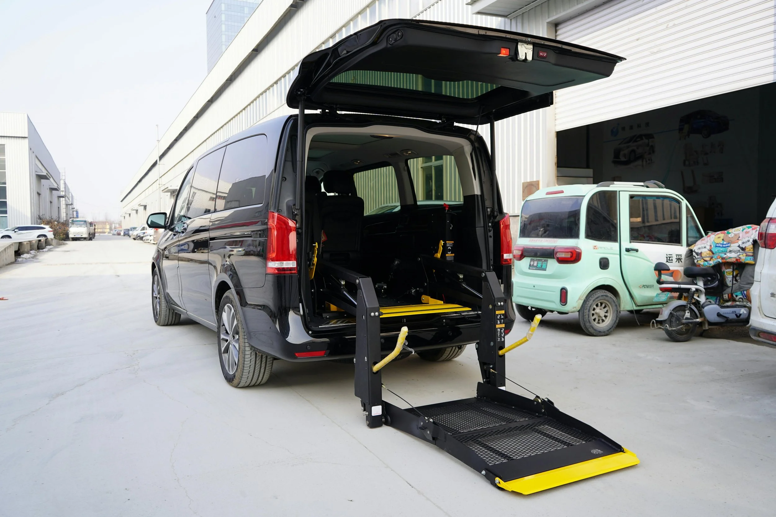 A black minivan with an open rear hatch showing a wheelchair lift attached to the back, parked outside a warehouse-like building with other vehicles nearby.