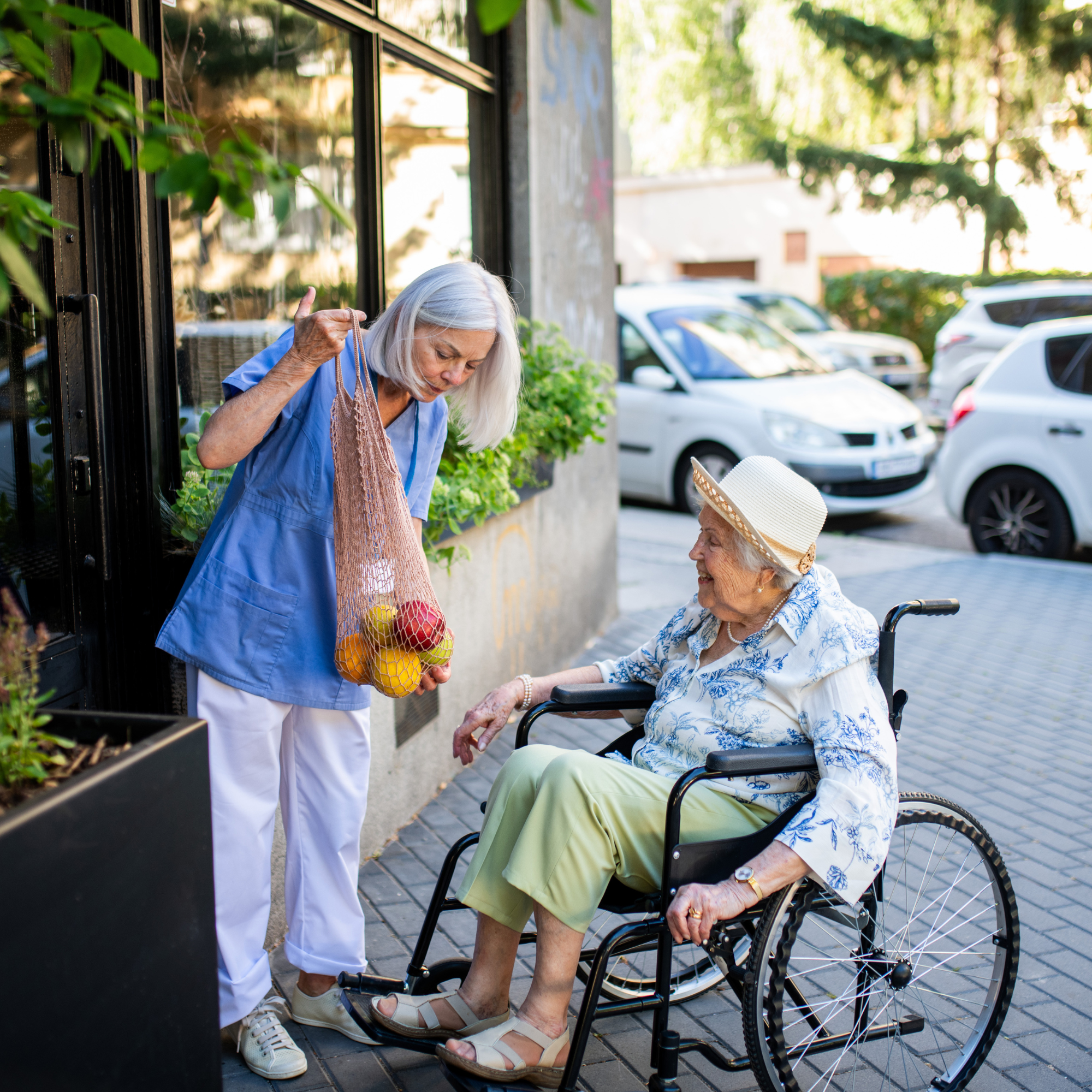 An elderly woman sitting in a wheelchair exchanging a handshake with a woman wearing a blue shirt and tan sandals, outside near parked cars, with greenery and a building in the background.