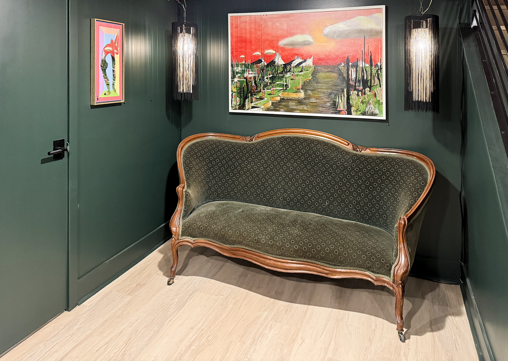 An antique green velvet loveseat with wood trim in a room with dark green walls. Above the loveseat are two modern black pendant lights hanging from the ceiling. There are two colorful artworks on the wall, one large landscape painting and one smaller, vertical piece. The room has light wood flooring.