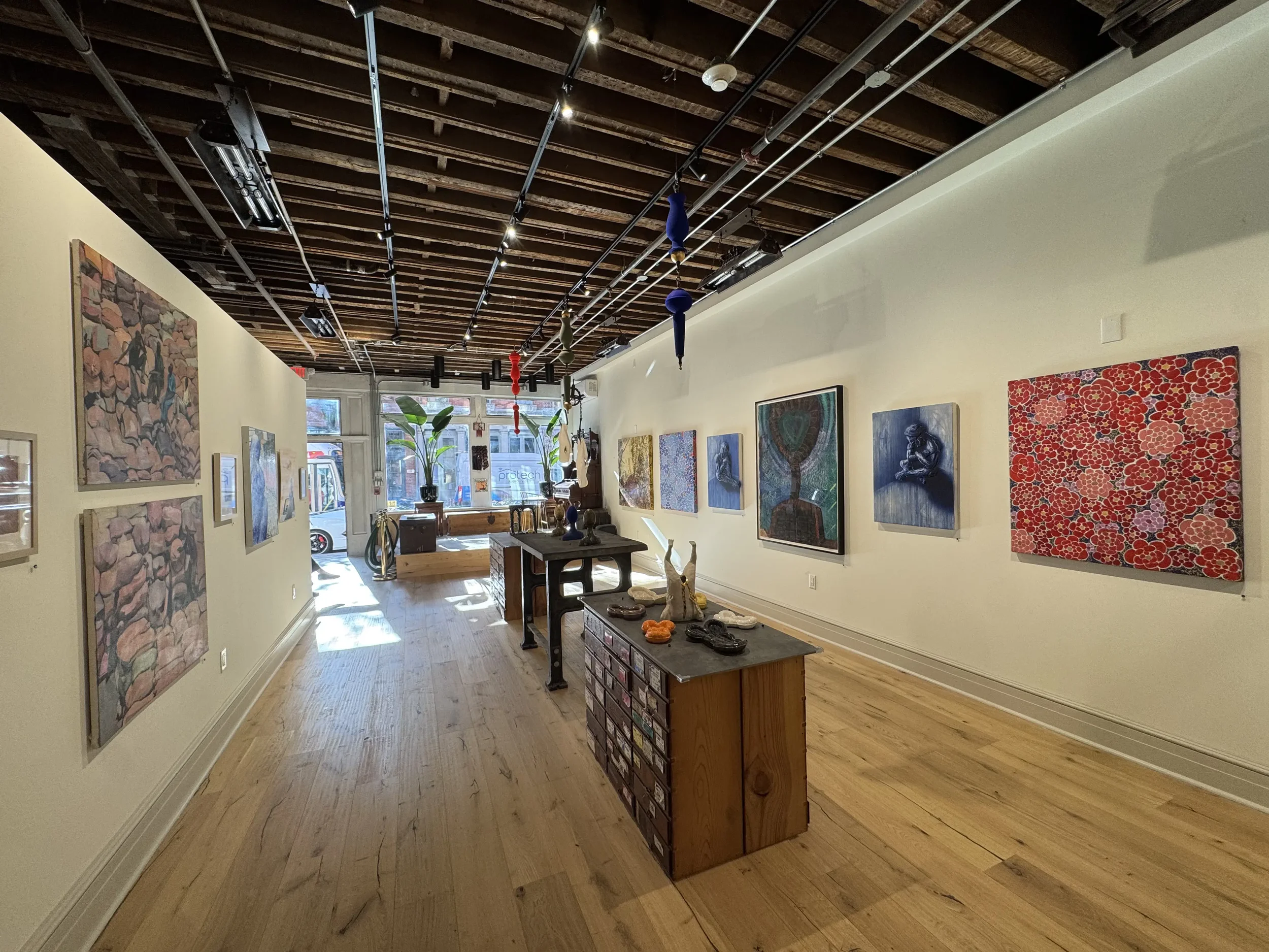 Interior of Curator Collective art gallery displaying paintings on the walls and sculptures on tables, with natural light coming through large windows and a wooden floor.