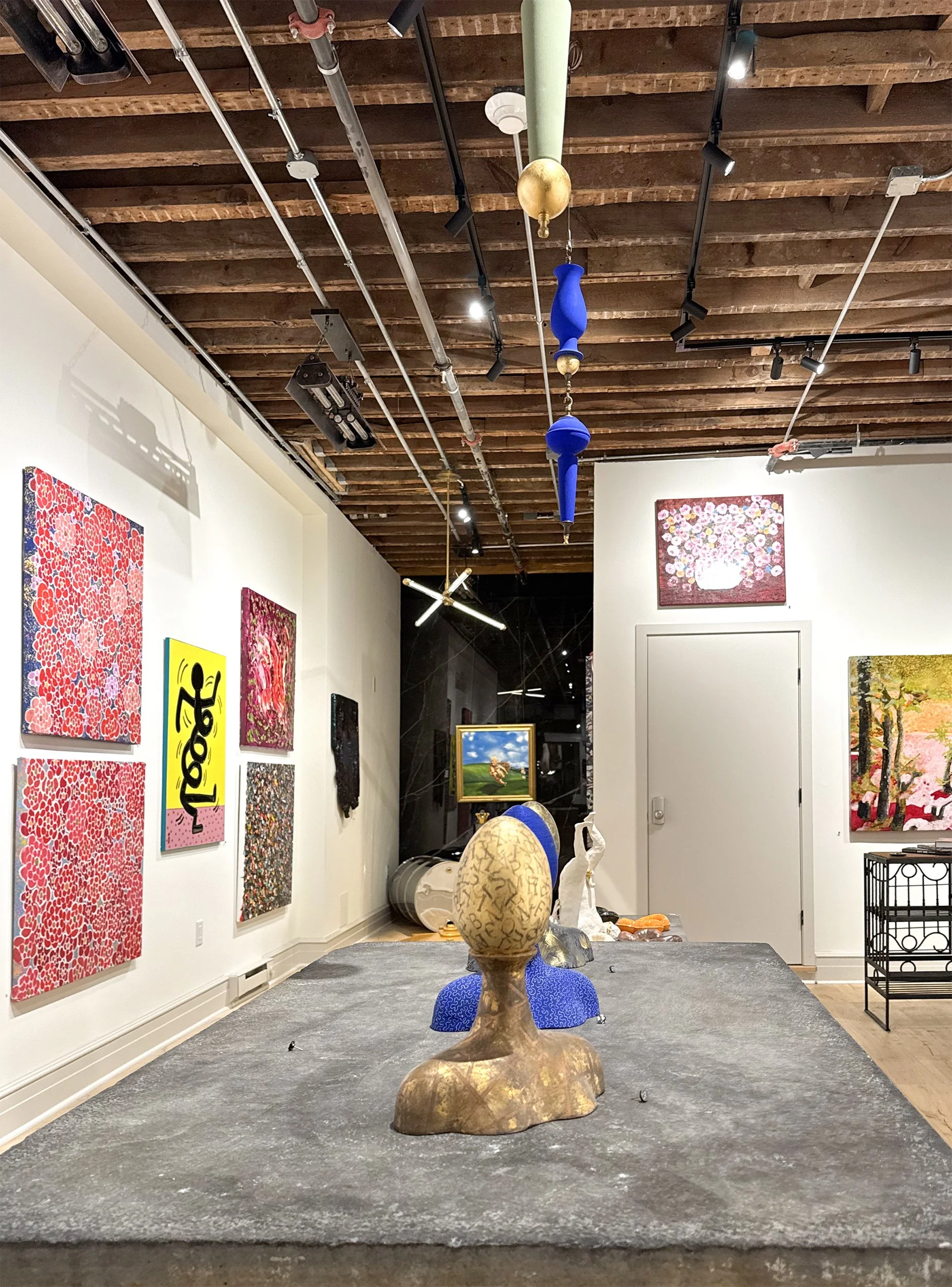 Art gallery with paintings on the wall, abstract sculptures on a gray carpeted floor, and a wooden ceiling with track lighting.