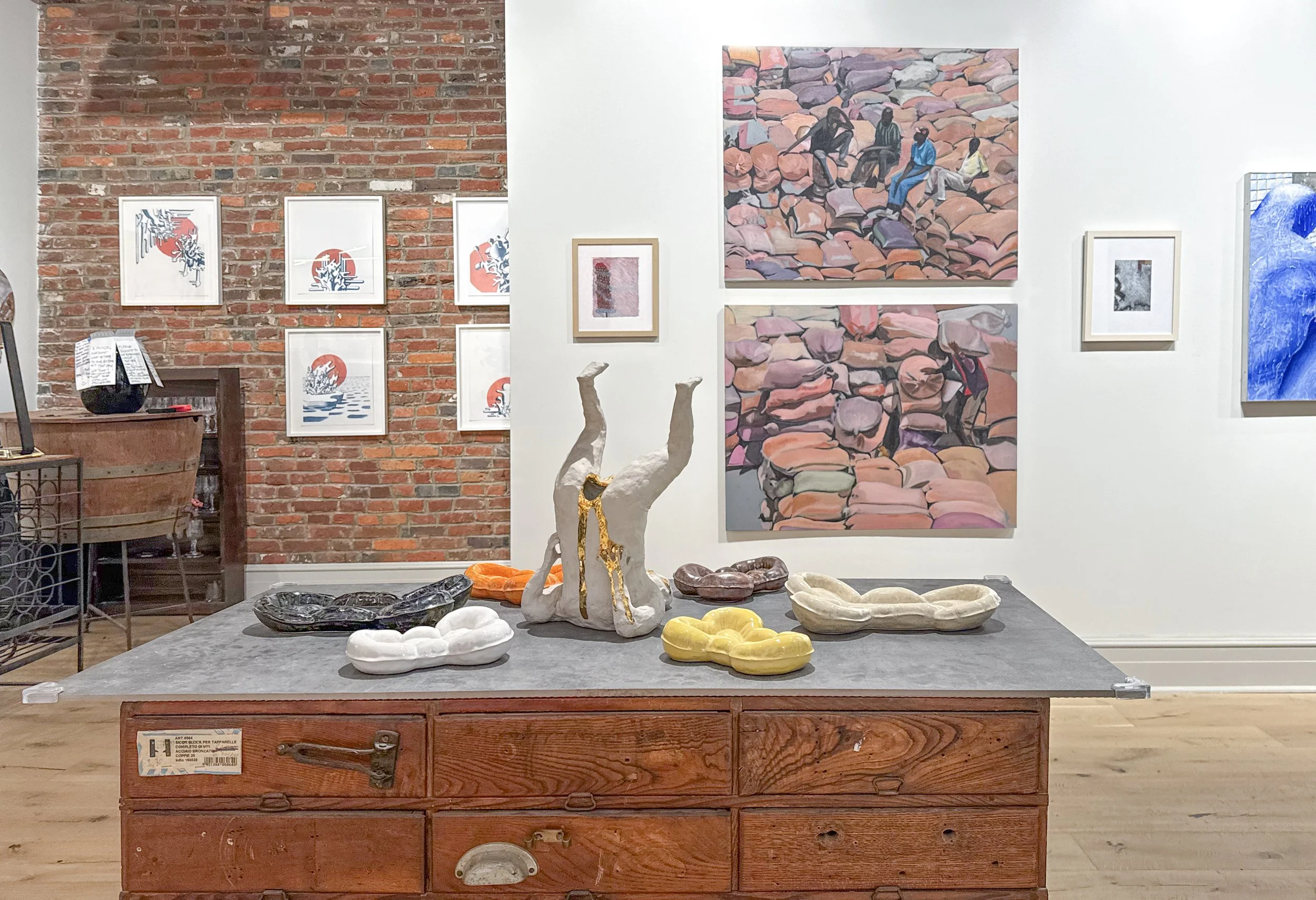 An art gallery with a brick wall and white wall, displaying various paintings and artworks. A wooden table in the foreground holds decorative objects, sculptures, and ceramic bowls. The artworks include paintings of rocks, abstract designs, and a landscape with blue tones.