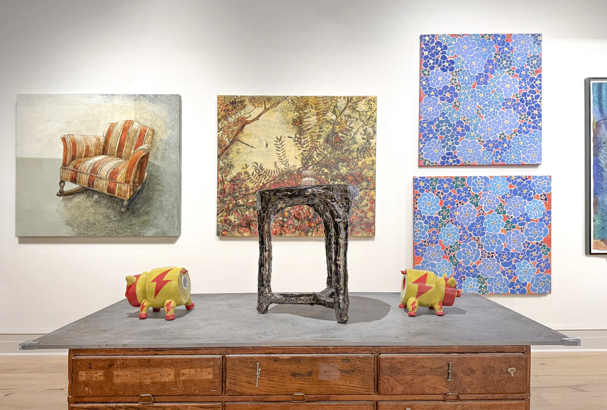 An art gallery exhibit features several pieces of artwork displayed on a white wall and a wooden platform. There are three paintings, including one of a striped upholstered armchair, a landscape with trees and flowers, and an abstract floral pattern in blue tones. In front of the display, there are carved wooden stools and two yellow decorative piggy banks with red lightning bolt designs.
