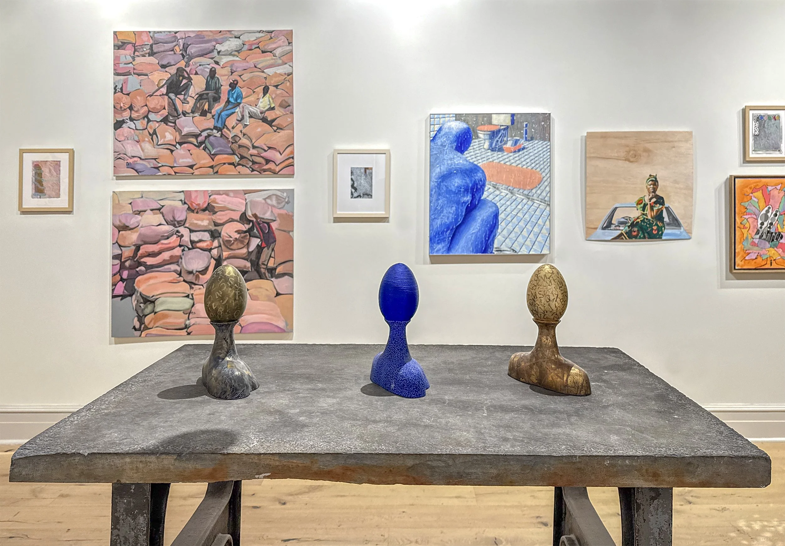 Art gallery with paintings on the wall and four ceramic or stone sculptures on a table in front.