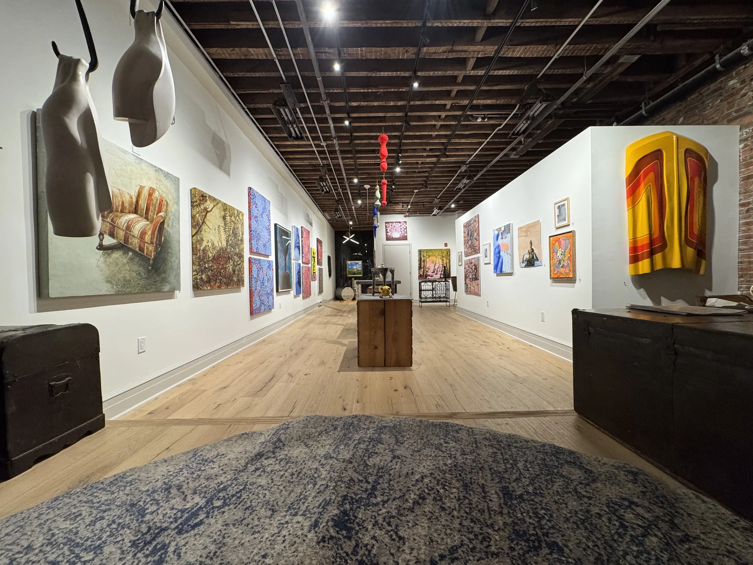 An art gallery with various paintings on white walls, showcasing a diverse range of art styles and subjects. The gallery features a wooden floor, a brick wall on the right, and a ceiling with exposed wooden beams and pipes. There are ceiling-mounted lights illuminating the artworks, and some hanging sculptures are visible on the left side of the image.