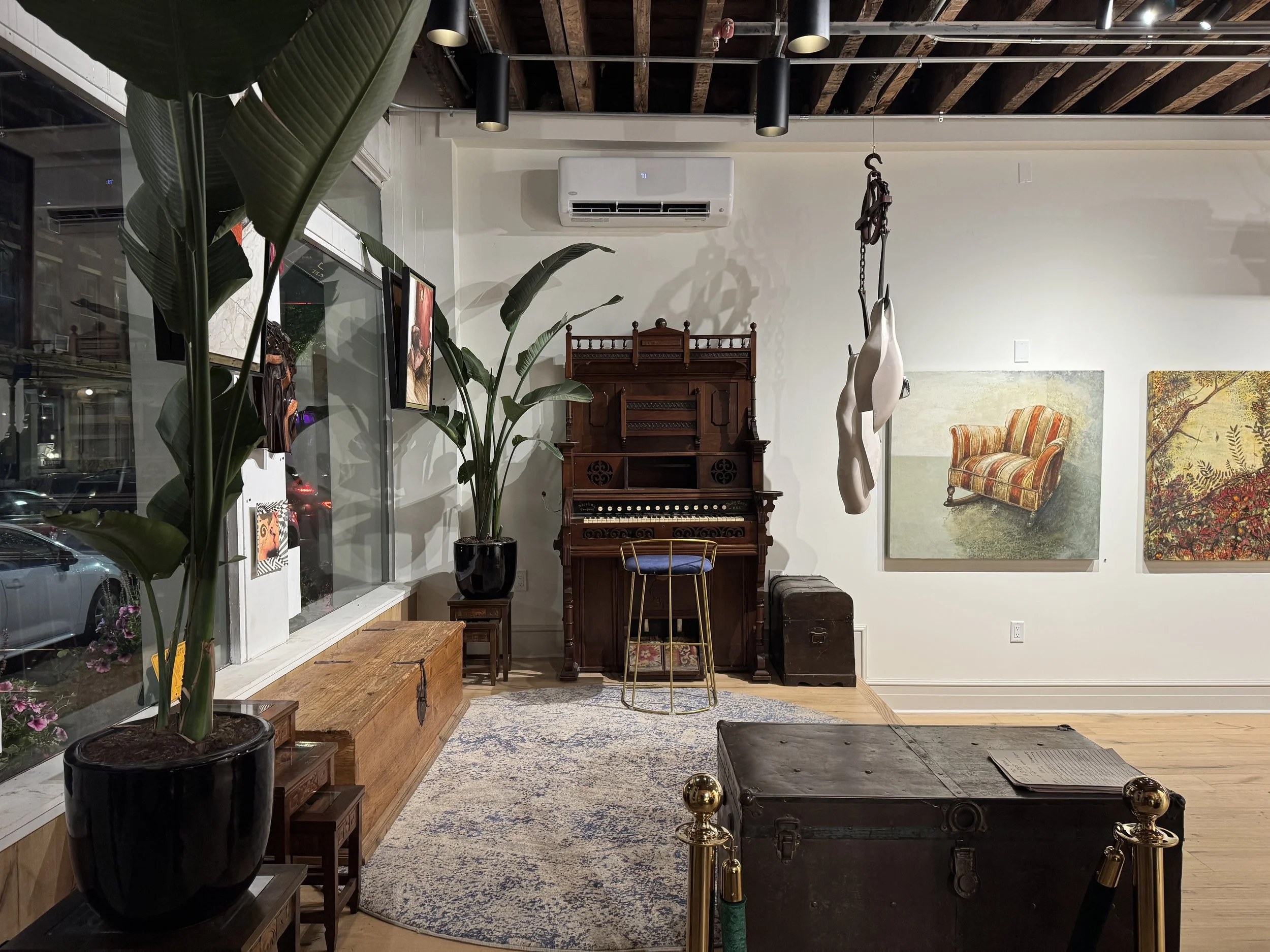 Interior of a modern art gallery with various paintings and sculptures on display, including a vintage wooden organ, a large abstract painting, and sculptures of hanging cloth resembling human figures. The space features an exposed wooden ceiling, large windows, and decorative rugs on the floor.
