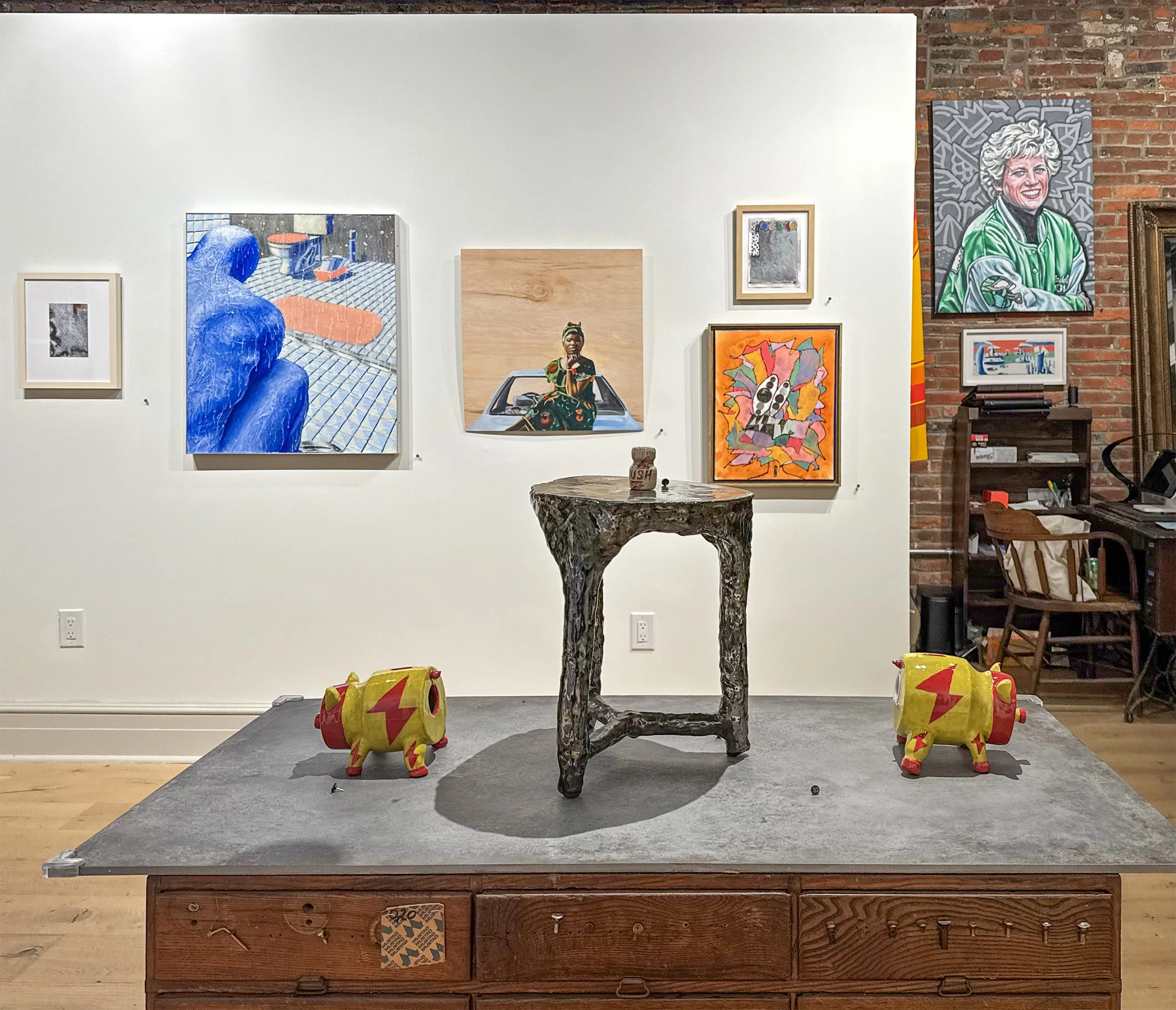 An art gallery display with a central carved wooden table, surrounded by four small yellow toy vehicles with red lightning bolt designs, mounted on a gray platform.