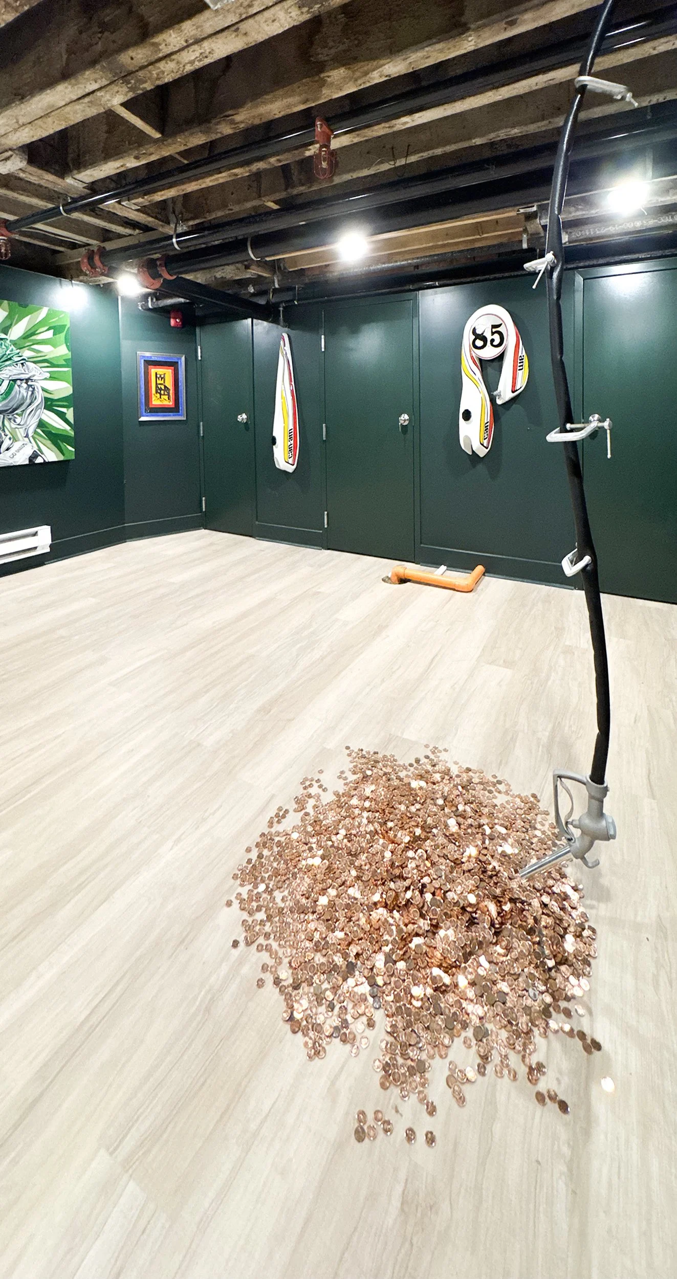A room with a pile of pennies on the floor, a mop, painted dark green walls with framed artwork, and ceiling with exposed wooden beams and pipes.