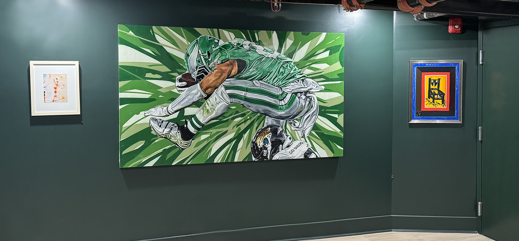 Painting of a football player wearing a green uniform and helmet, crouched and holding a football, with a background of green leaves, displayed on a dark green wall in an art gallery.