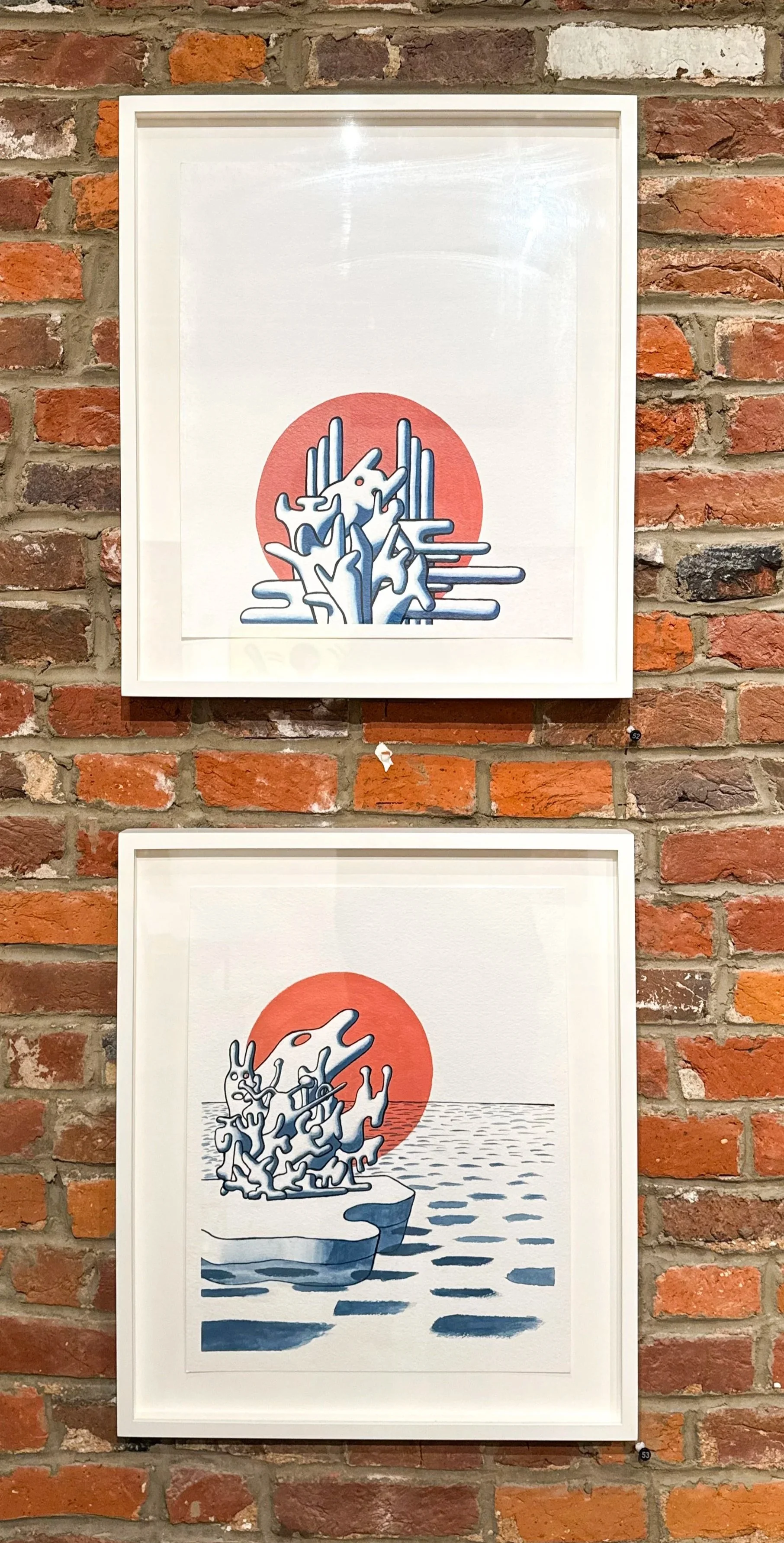 Two framed abstract art prints are hung on a brick wall, each featuring stylized trees with a red sun or moon in the background.