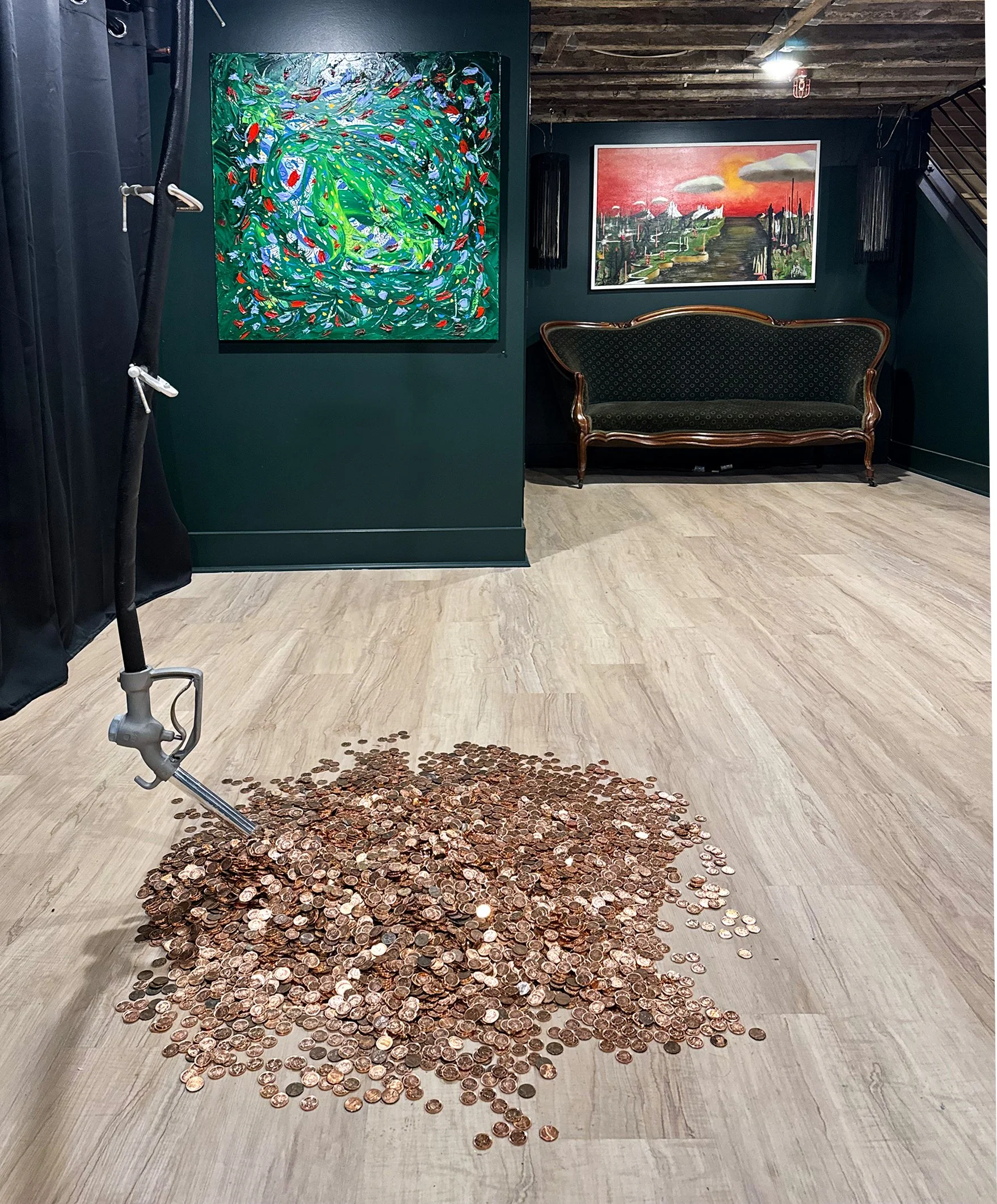 A collection of pennies spread out on a light-colored wooden floor, with some poured from a container, in front of a dark green wall with two paintings. The painting on the left features abstract swirls of green, blue, red, and white, and the painting on the right depicts a landscape with a red sunset sky and dark silhouettes of trees and structures.
