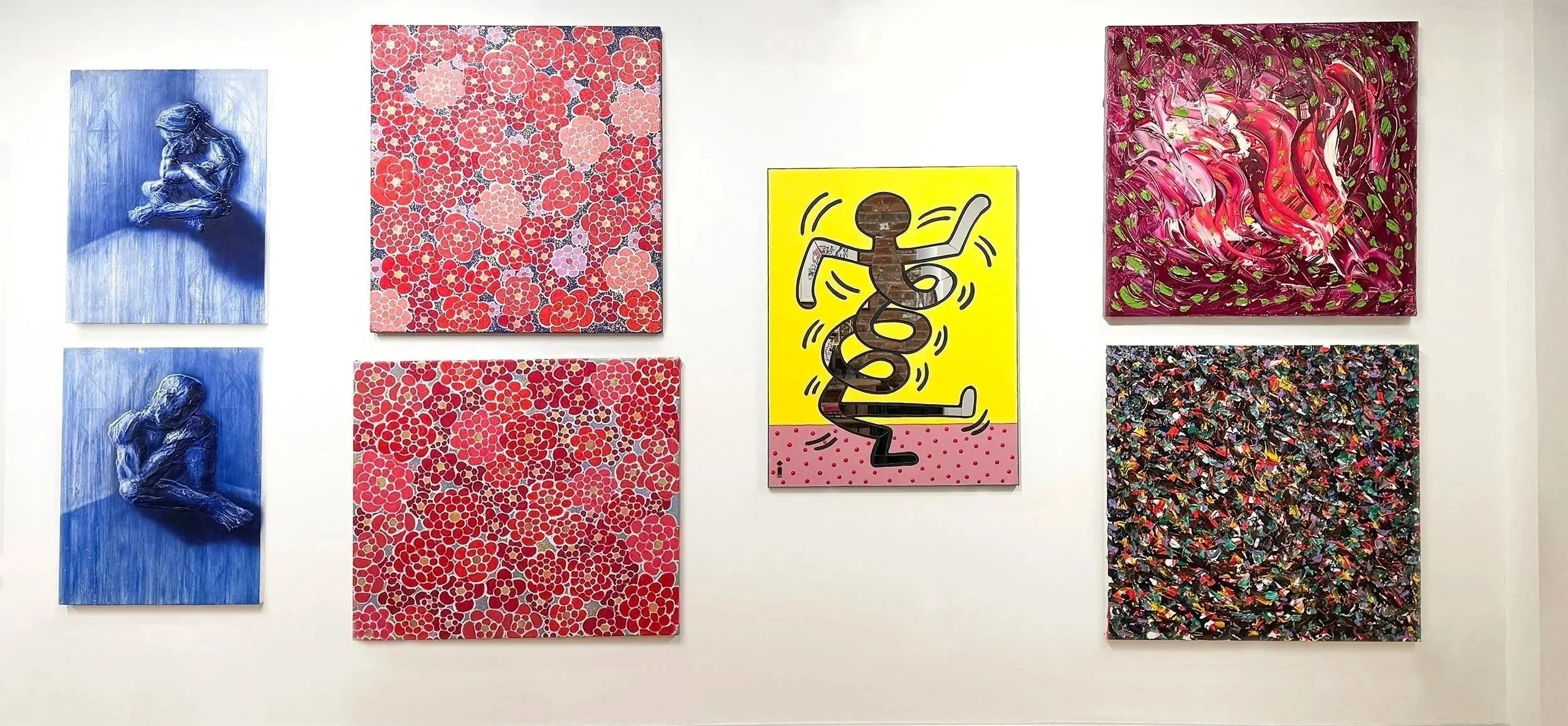 An art display on a white wall featuring eight paintings arranged in two rows. The top row includes three paintings: a blue monochrome artwork of a seated person, a floral pattern with pink and red flowers, and an abstract piece with swirling pink, white, and green brushstrokes. The bottom row includes two paintings: another blue monochrome artwork of a seated person and a densely textured multicolored abstract piece. In the center is a bright yellow painting with a cartoonish figure and the word 'good' written in a playful font.