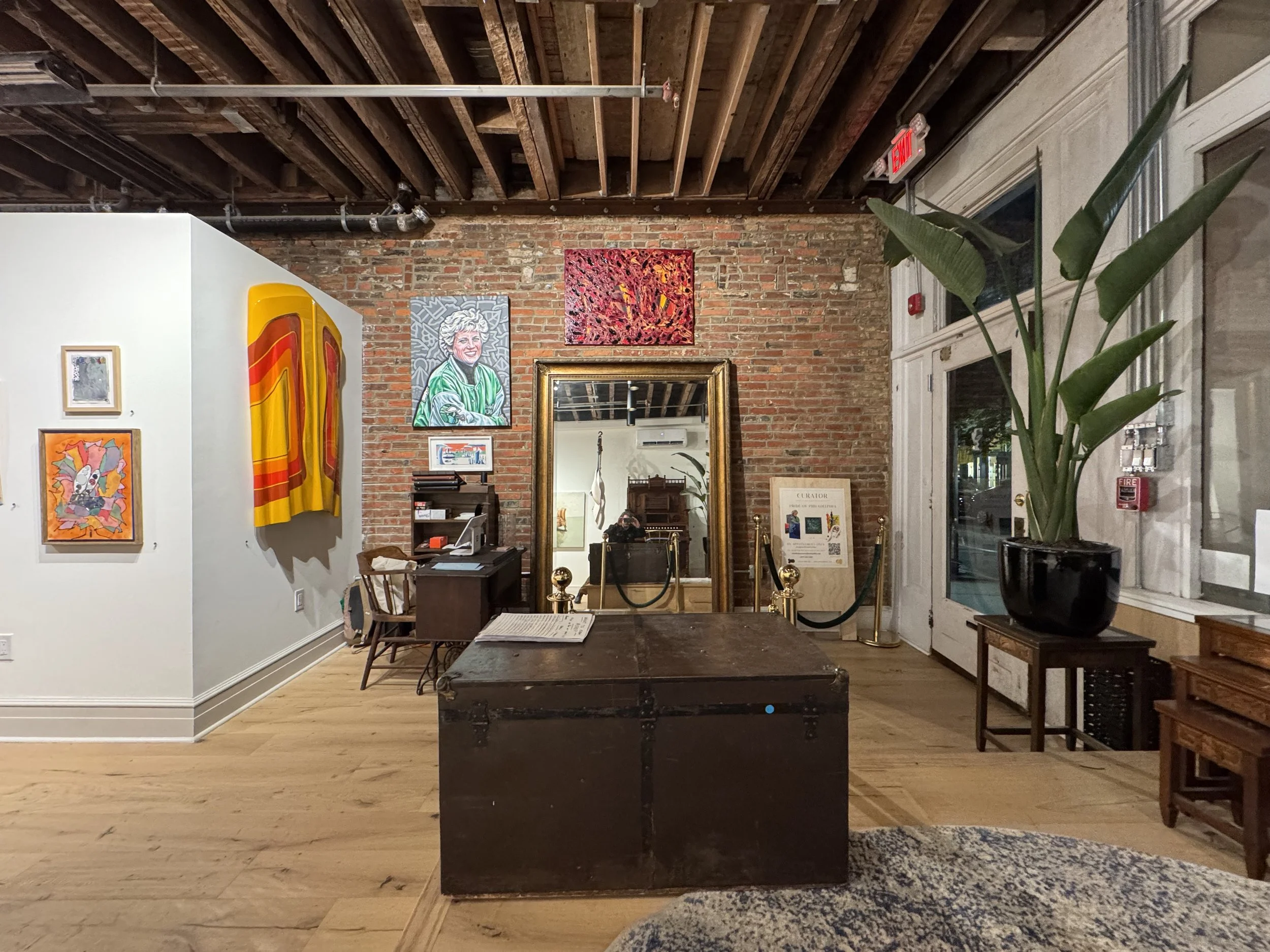 An interior art gallery with exposed brick walls and a wooden ceiling. Several colorful paintings are displayed on the walls, a tall green plant is in a black pot, and a large mirror is mounted on one wall. There is a wooden table with chairs and various framed artworks and informational displays around the space.
