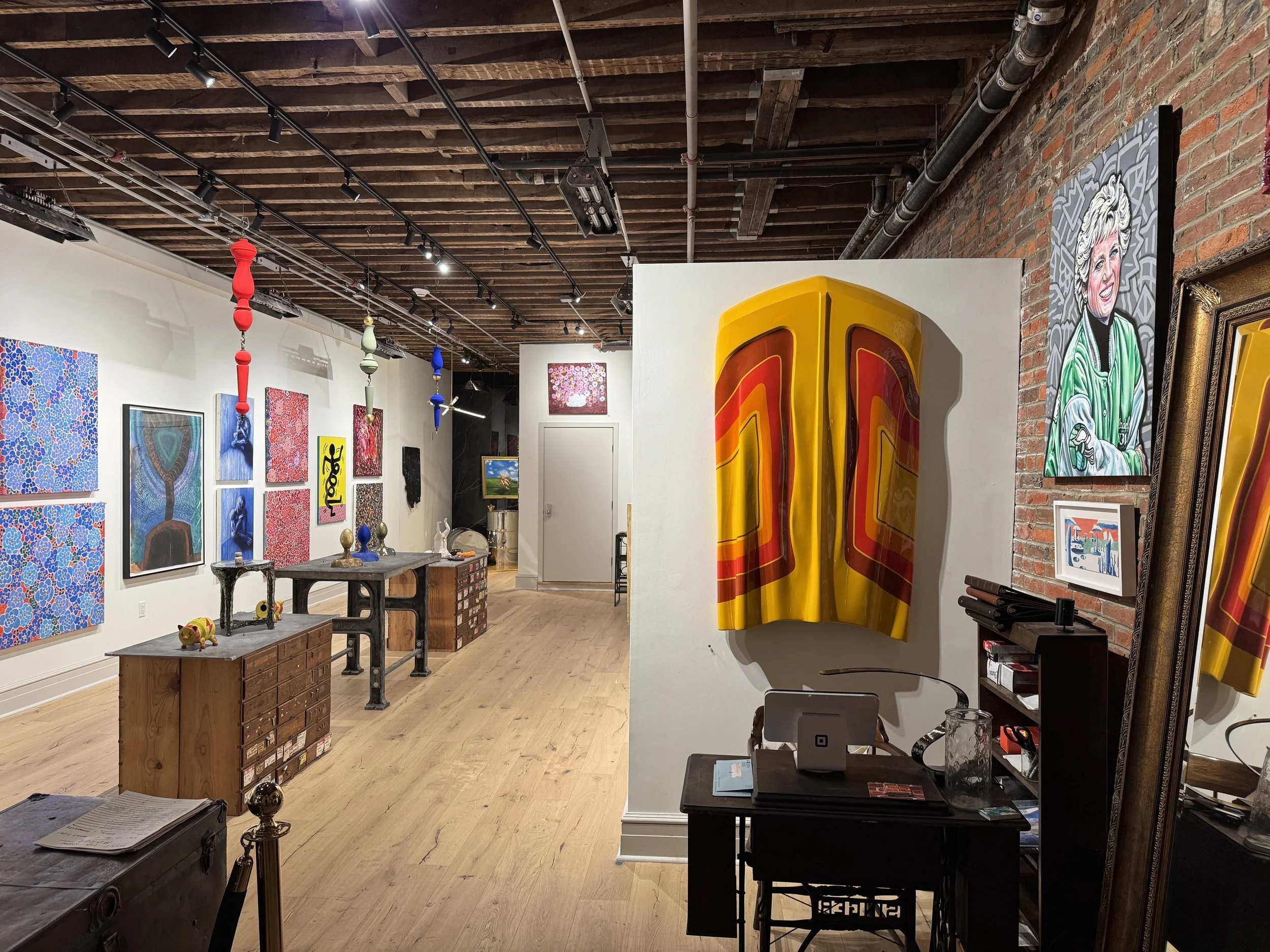 Art gallery interior with paintings on white and red brick walls, colorful sculptures, and wooden flooring.