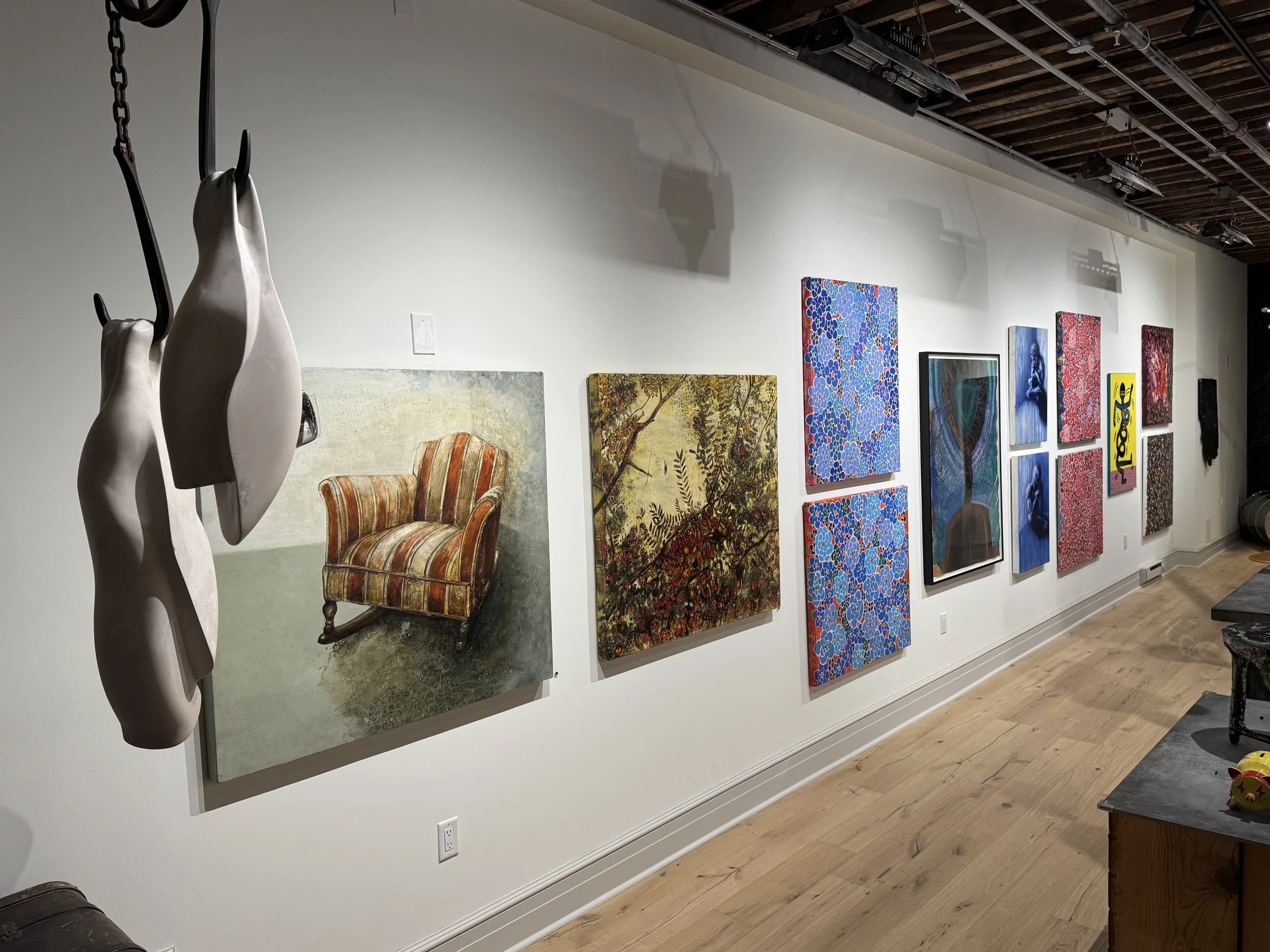 An art gallery wall with various paintings, including floral, portrait, and abstract art. In the foreground, there are two white abstract sculptures hanging from a chain. A vintage armchair with striped upholstery and a small table with a yellow toy are also visible.