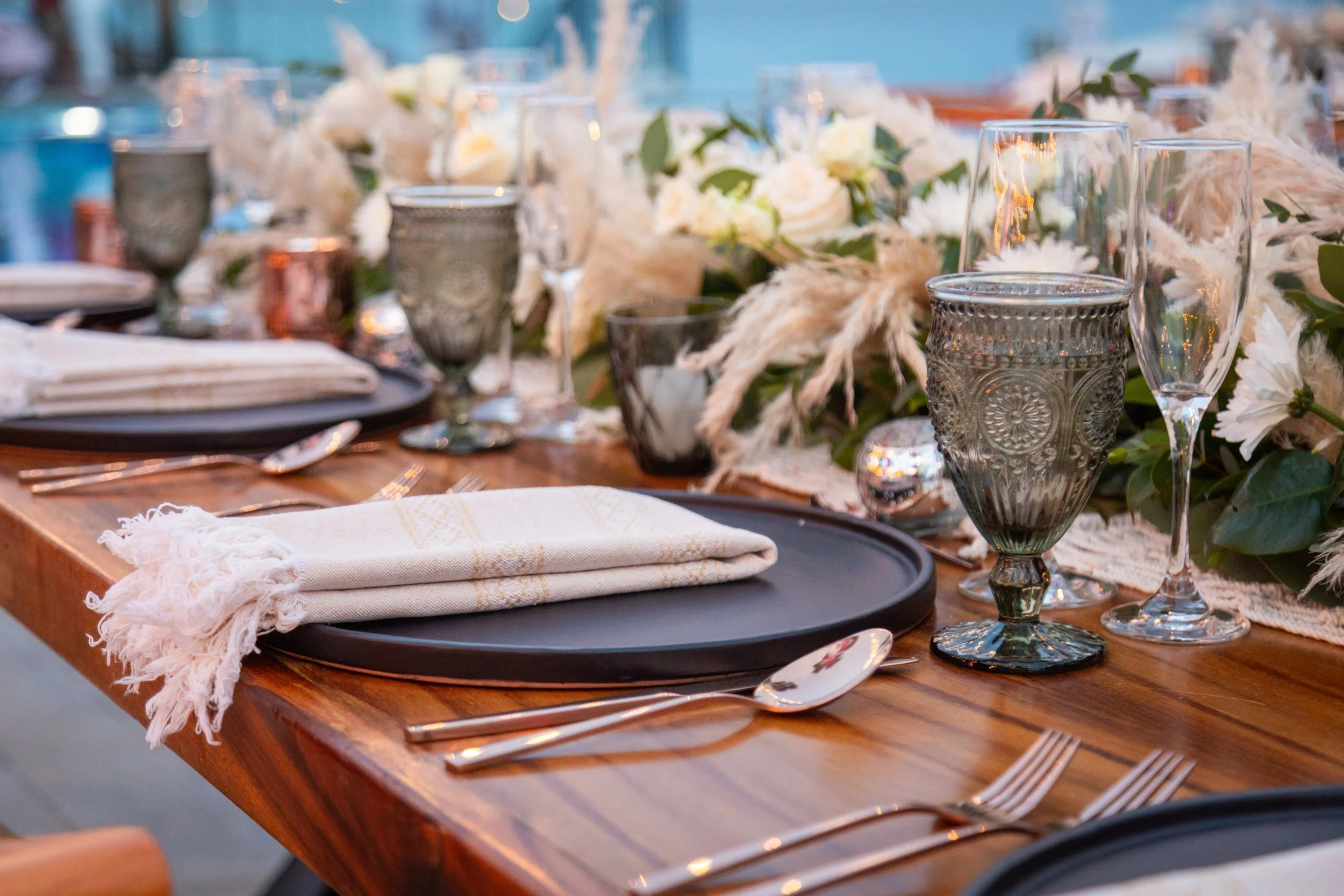 A close-up of an elegant dining table set with black charger plates, white cloth napkins, and silverware. The table features a centerpiece with white flowers and greenery, with decorative glassware and candles.