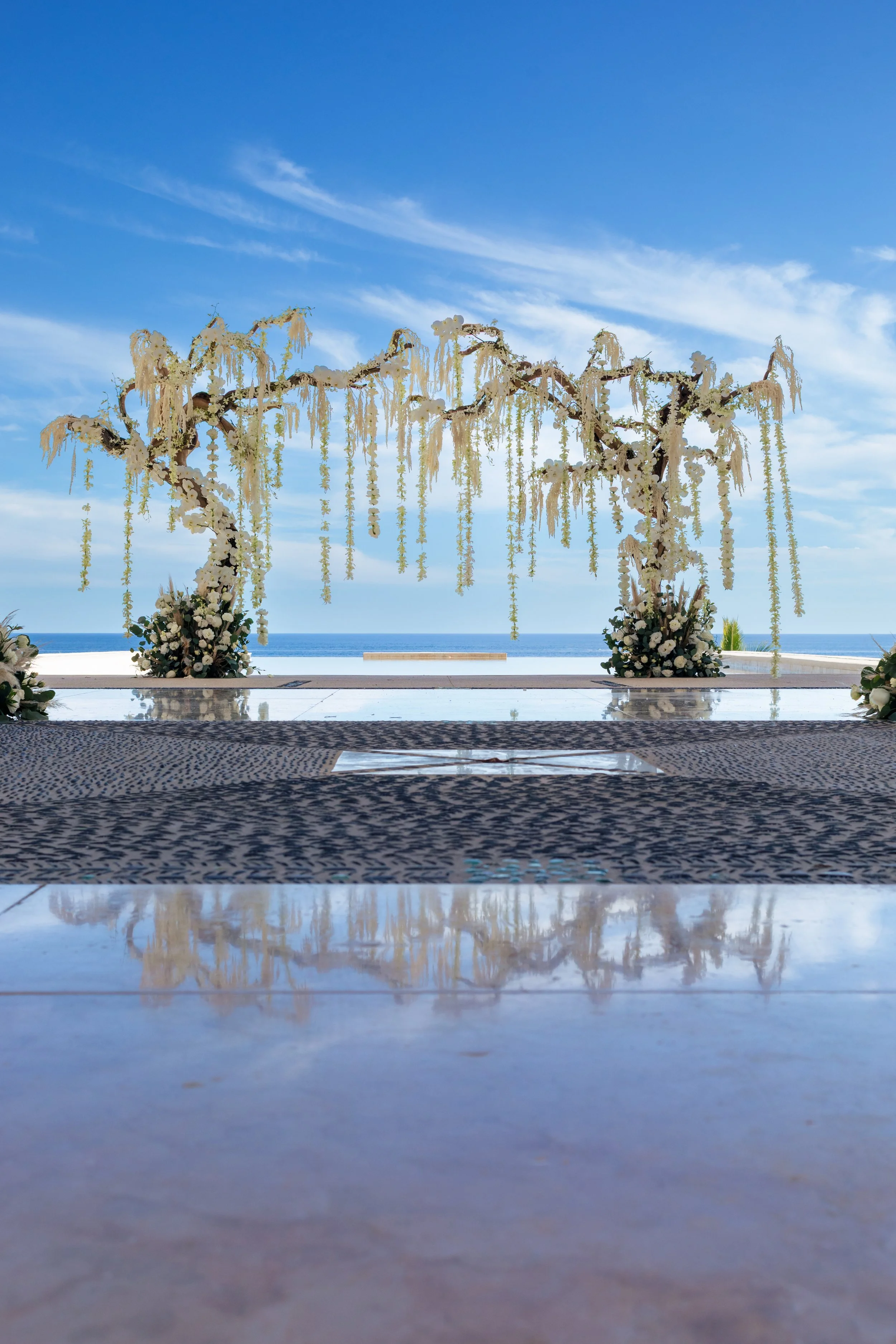 Oceanfront wedding ceremony decor in Los Cabos featuring a floral arch, white flowers, hanging greenery, and a romantic coastal ceremony setup by I DO Weddings Los Cabos.