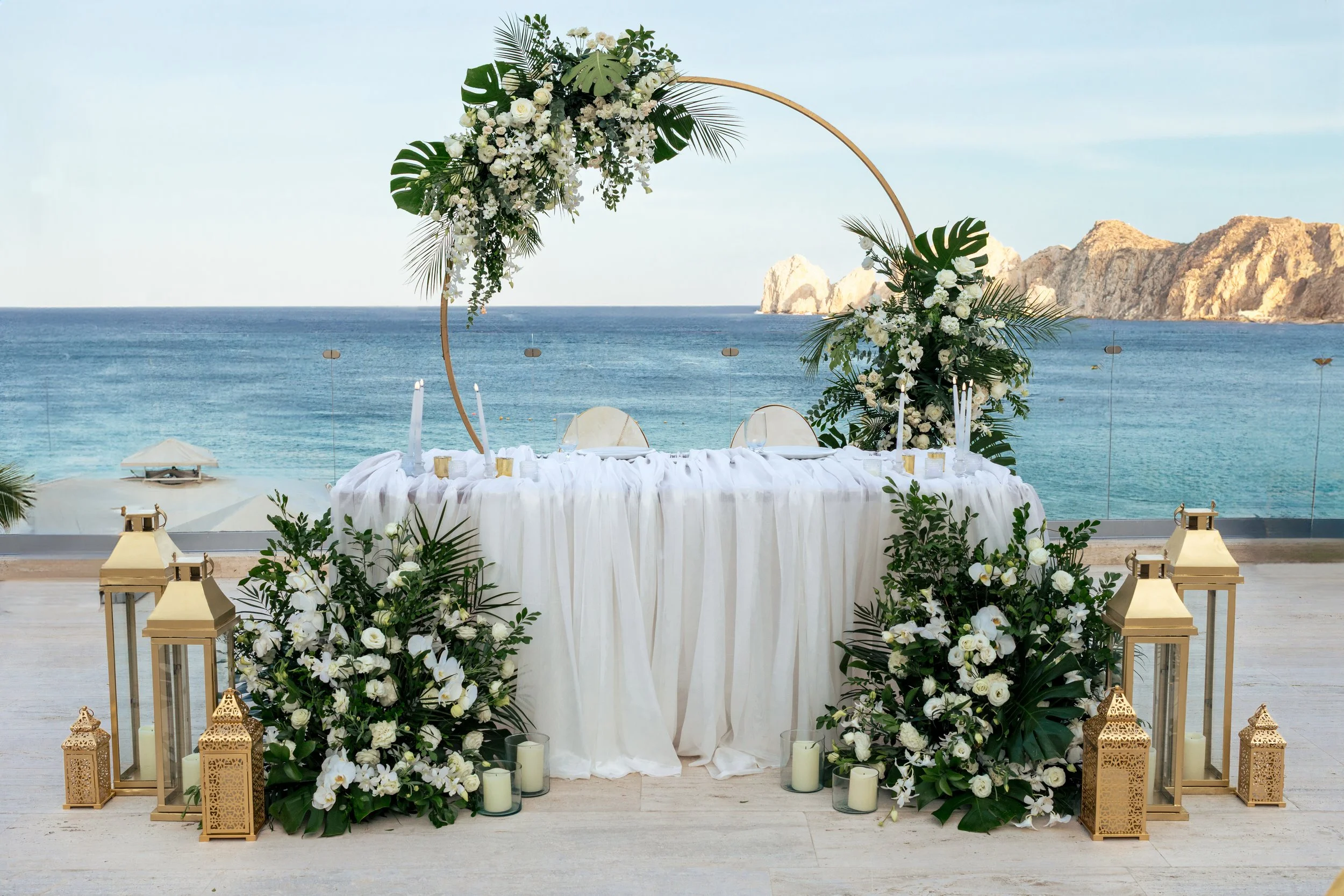 A wedding altar decorated with white flowers, green leaves, and a large circular wooden arch, overlooking the ocean with rocky cliffs in the background.