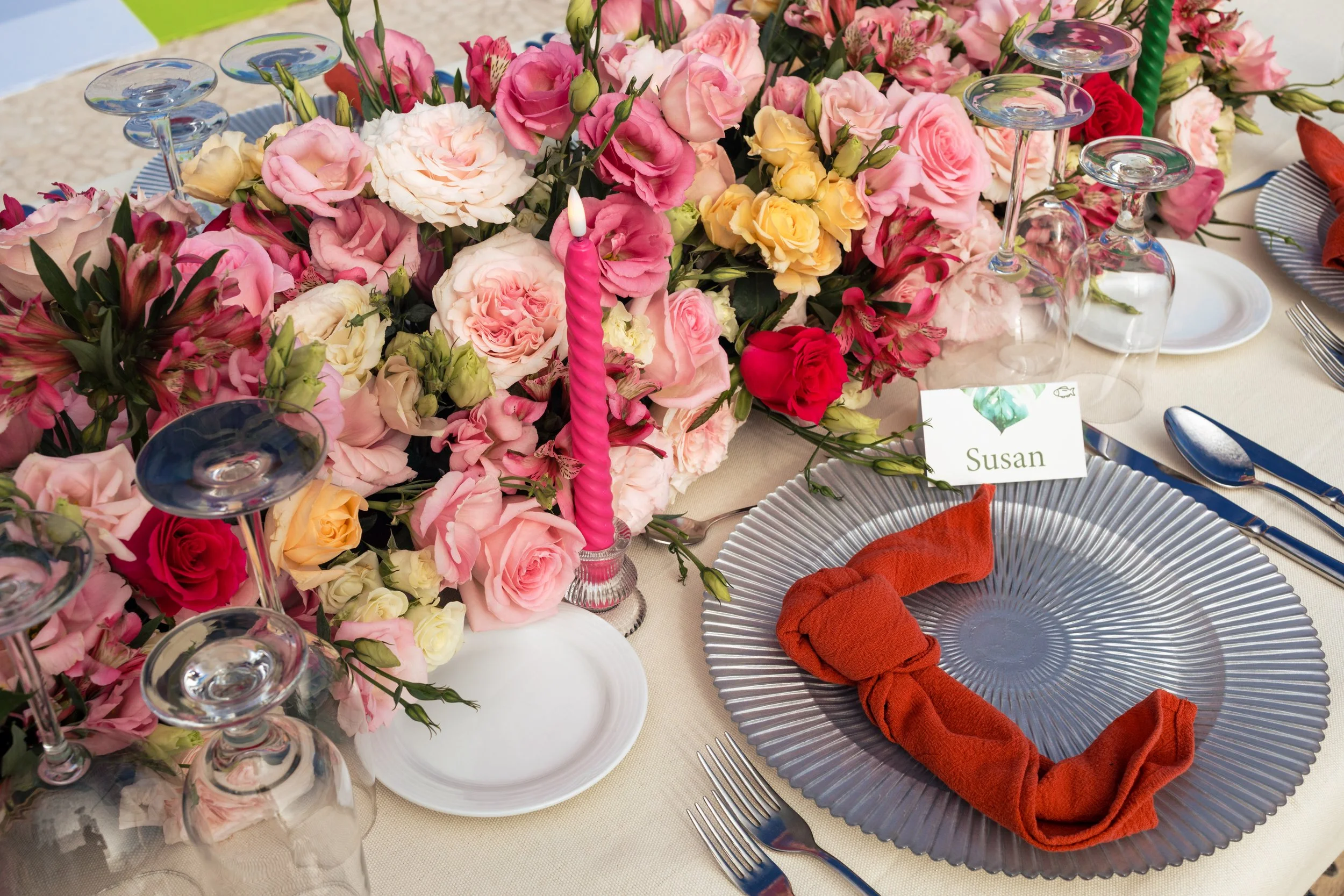 A table decorated with a large floral arrangement of pink, white, and yellow roses and lisianthus, with upside-down wine glasses, a pink twisted birthday candle, and place settings including a clear textured plate with an orange cloth napkin and silverware.