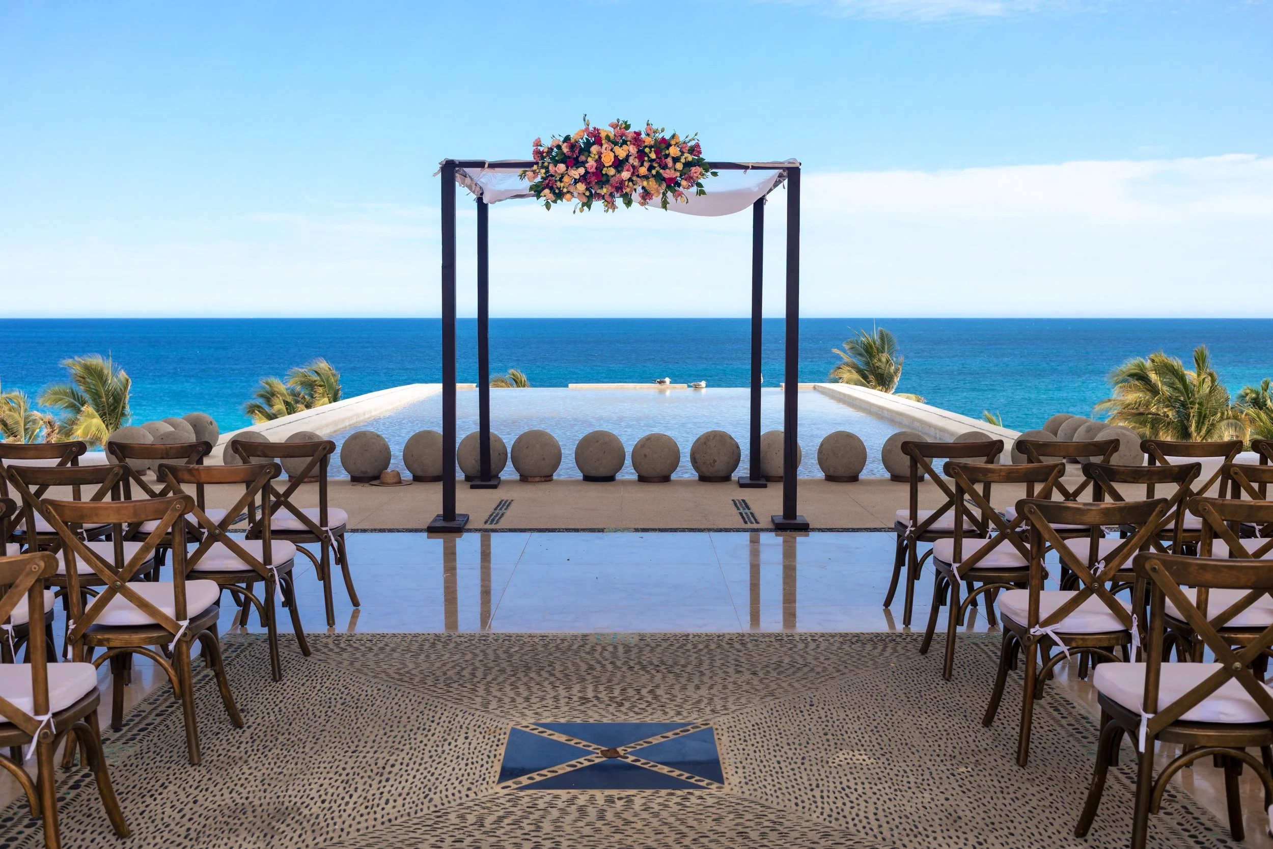 A wedding ceremony setup overlooking the ocean, with a floral arch on a platform and wooden chairs arranged on a patterned stone floor.