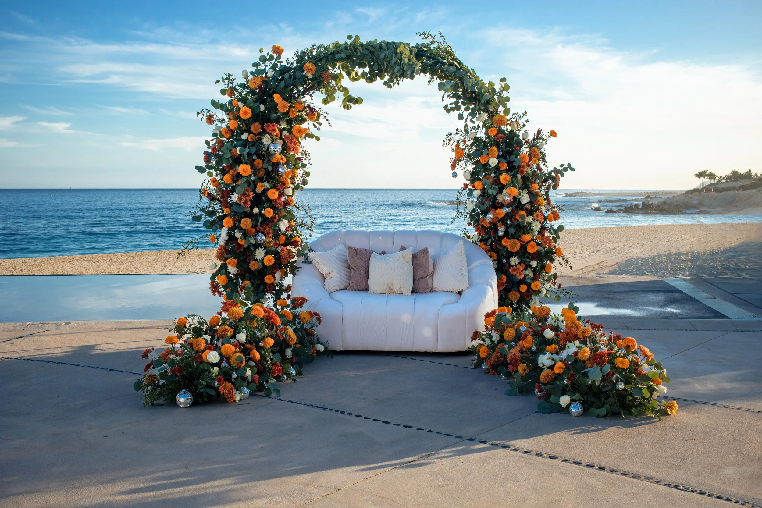 A white sofa decorated with pillows sits in front of an elaborate floral arch on a beach, with the ocean and sky in the background.