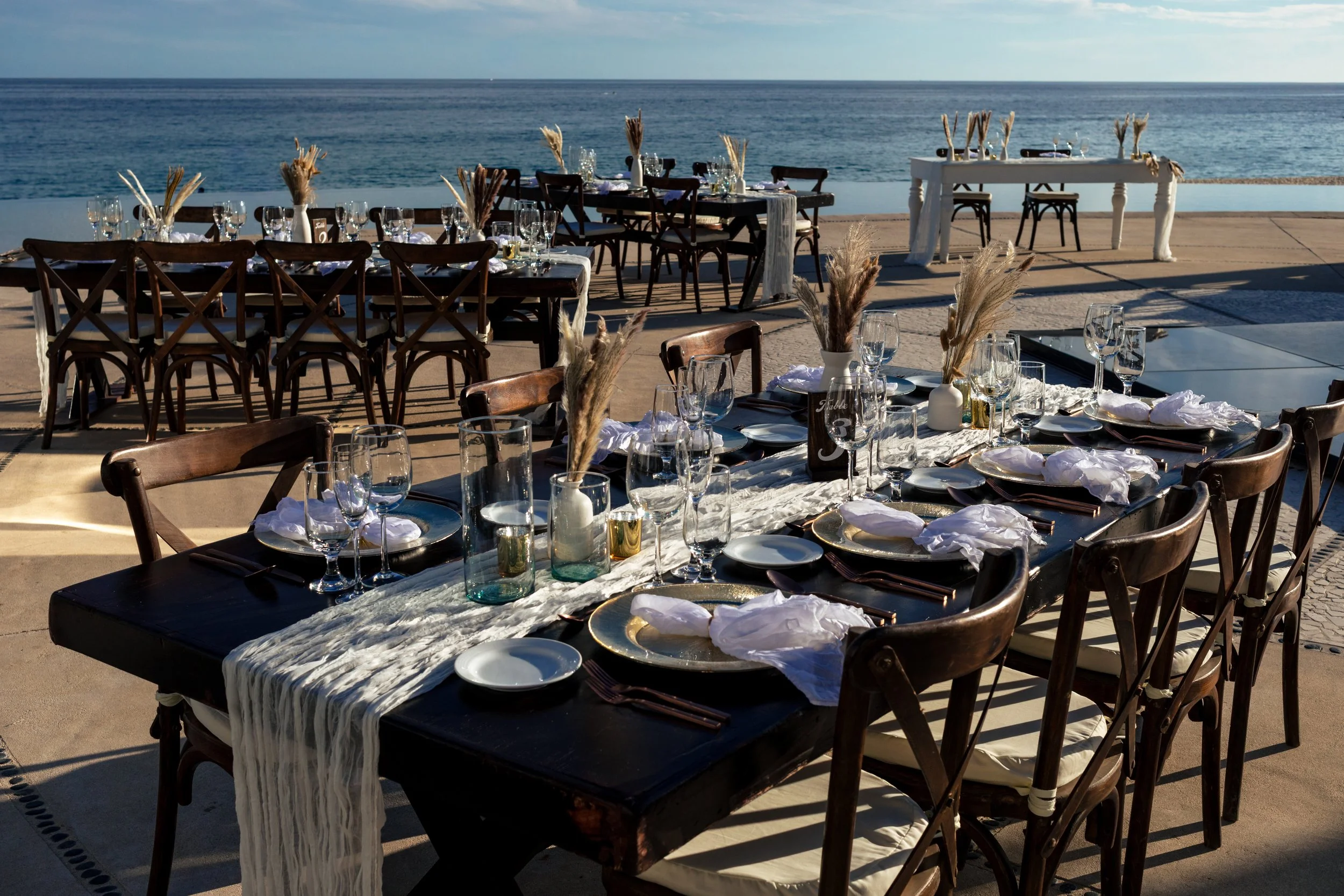 Outdoor wedding reception in Los Cabos with wooden chairs, decorated tables, tabletop details, and oceanfront event styling by I DO Weddings Los Cabos.