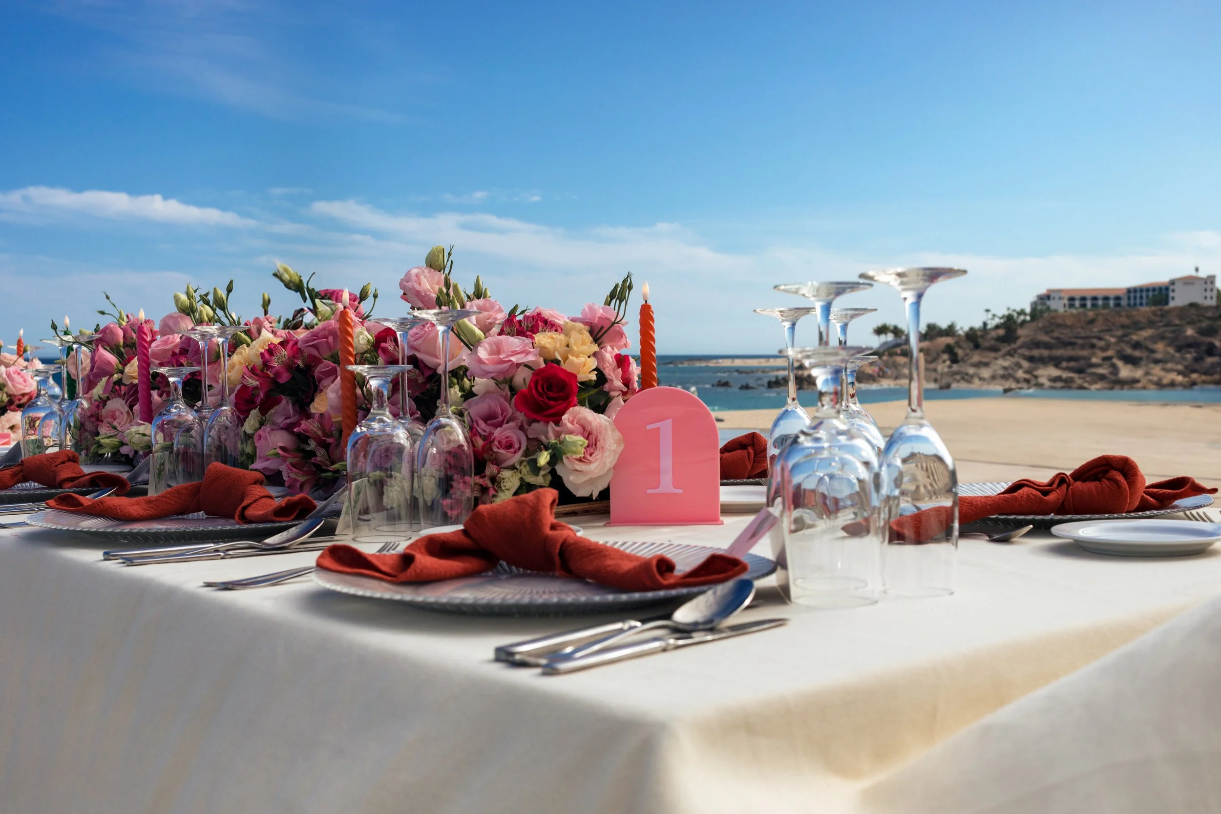 Beachside outdoor dining table decorated with pink and red flowers, candles, glassware, and orange napkins, with a coastal landscape and clear blue sky in the background.