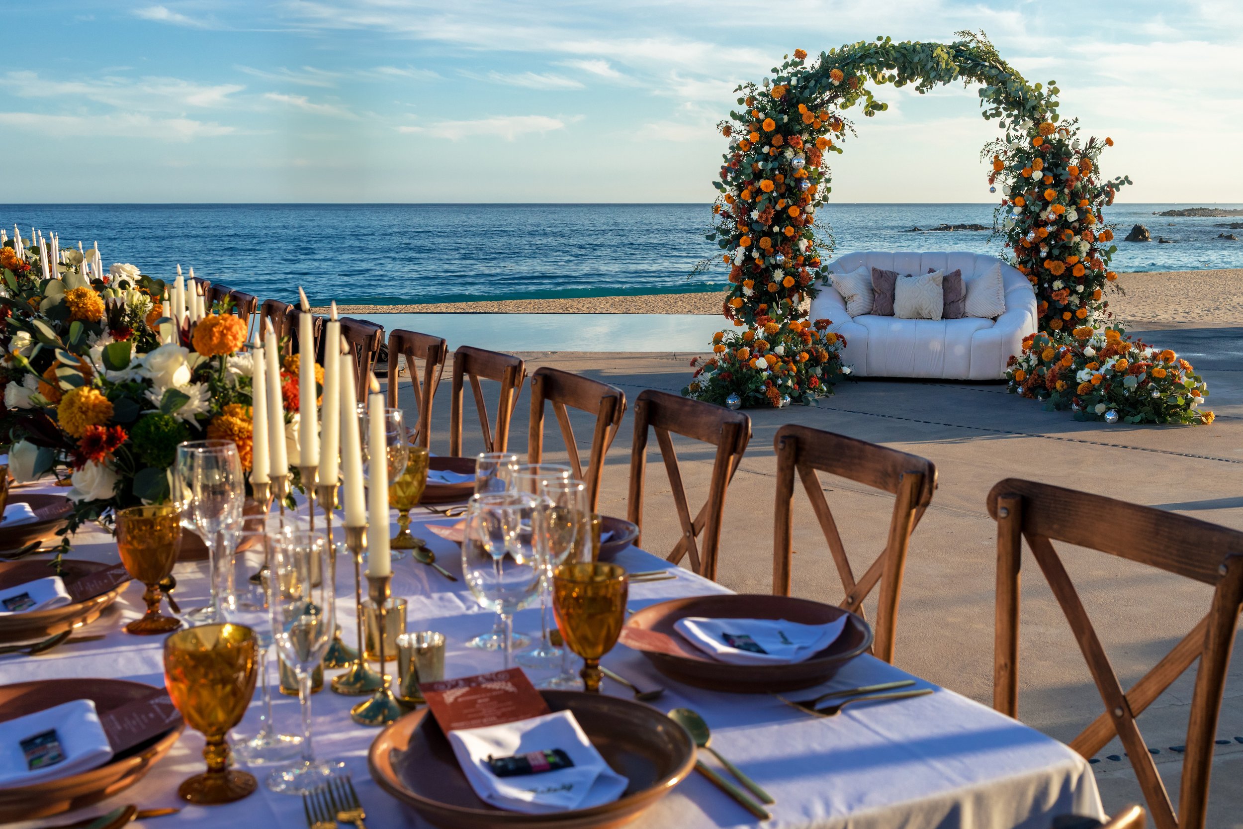 Beachside wedding setup in Los Cabos with floral arch, lounge seating, candles, tabletop details, and oceanfront decor by I DO Weddings Los Cabos.