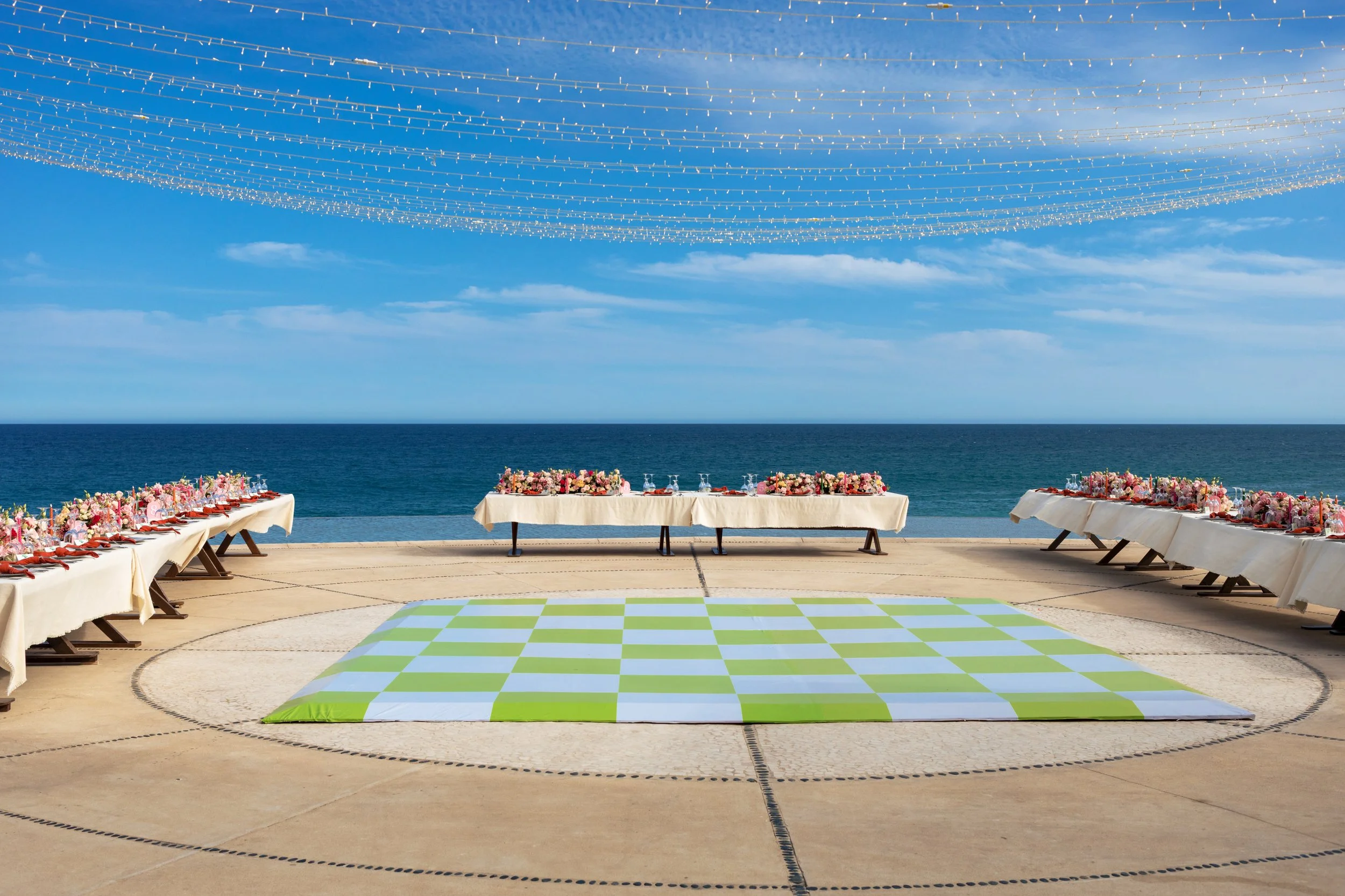 A beachside event setup with tables decorated with pink and white flowers and table settings, overlooking the ocean. The sky is bright with clouds, and decorative string lights are hung across the sky above the tables, creating an elegant seaside celebration atmosphere.