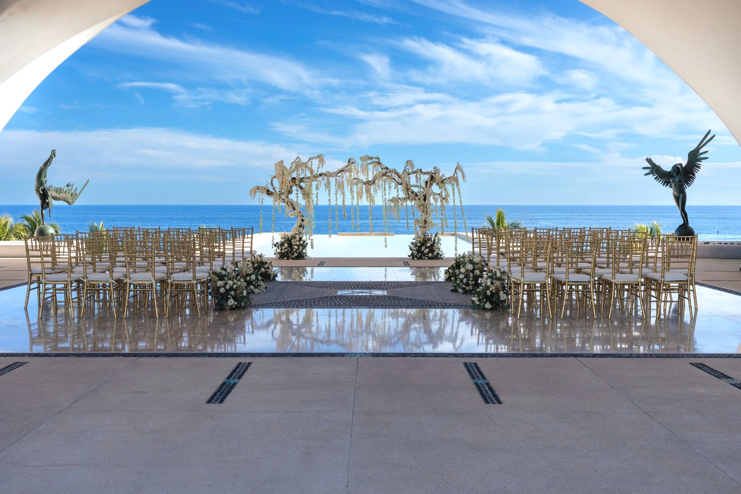 Oceanfront wedding ceremony setup in Los Cabos with floral ceremony structure, gold chairs, aisle decor, and coastal views by I DO Weddings Los Cabos.