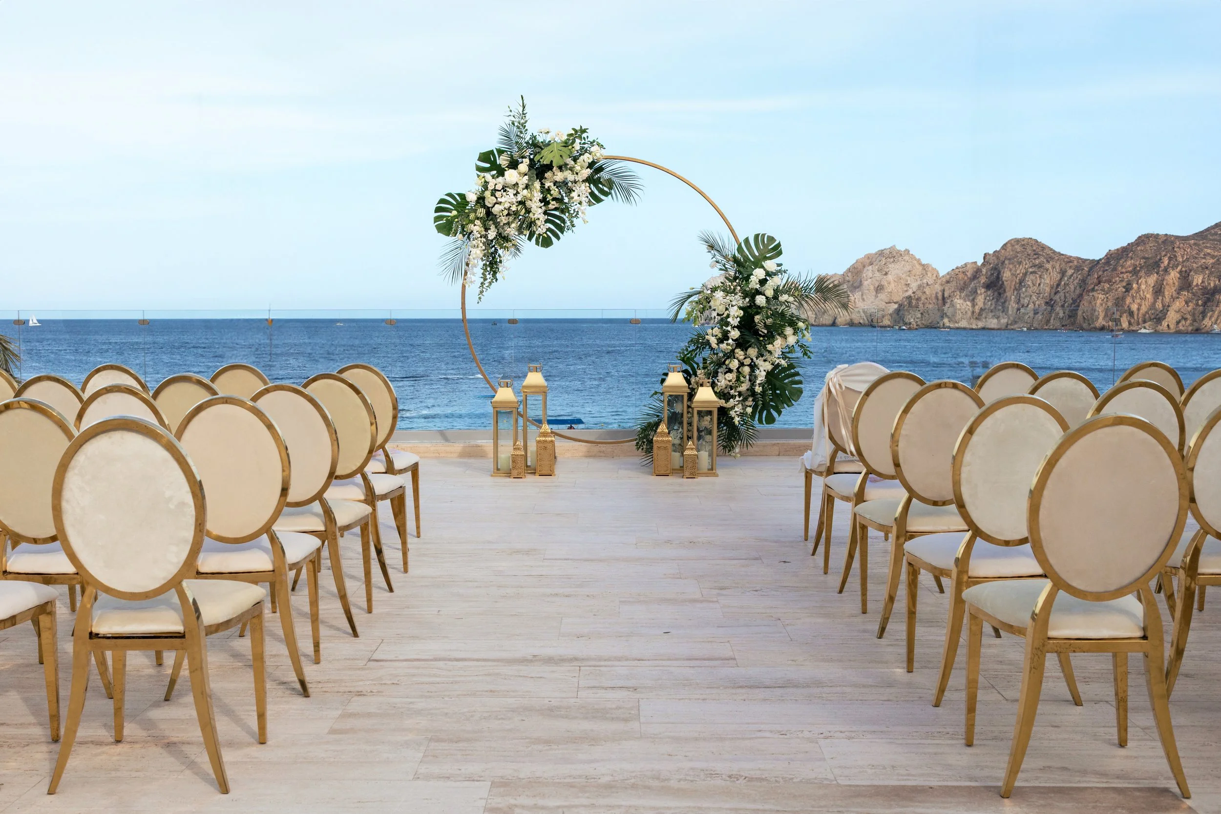 Outdoor wedding ceremony setup on a terrace overlooking the ocean with a circular floral altar and rows of white chairs.