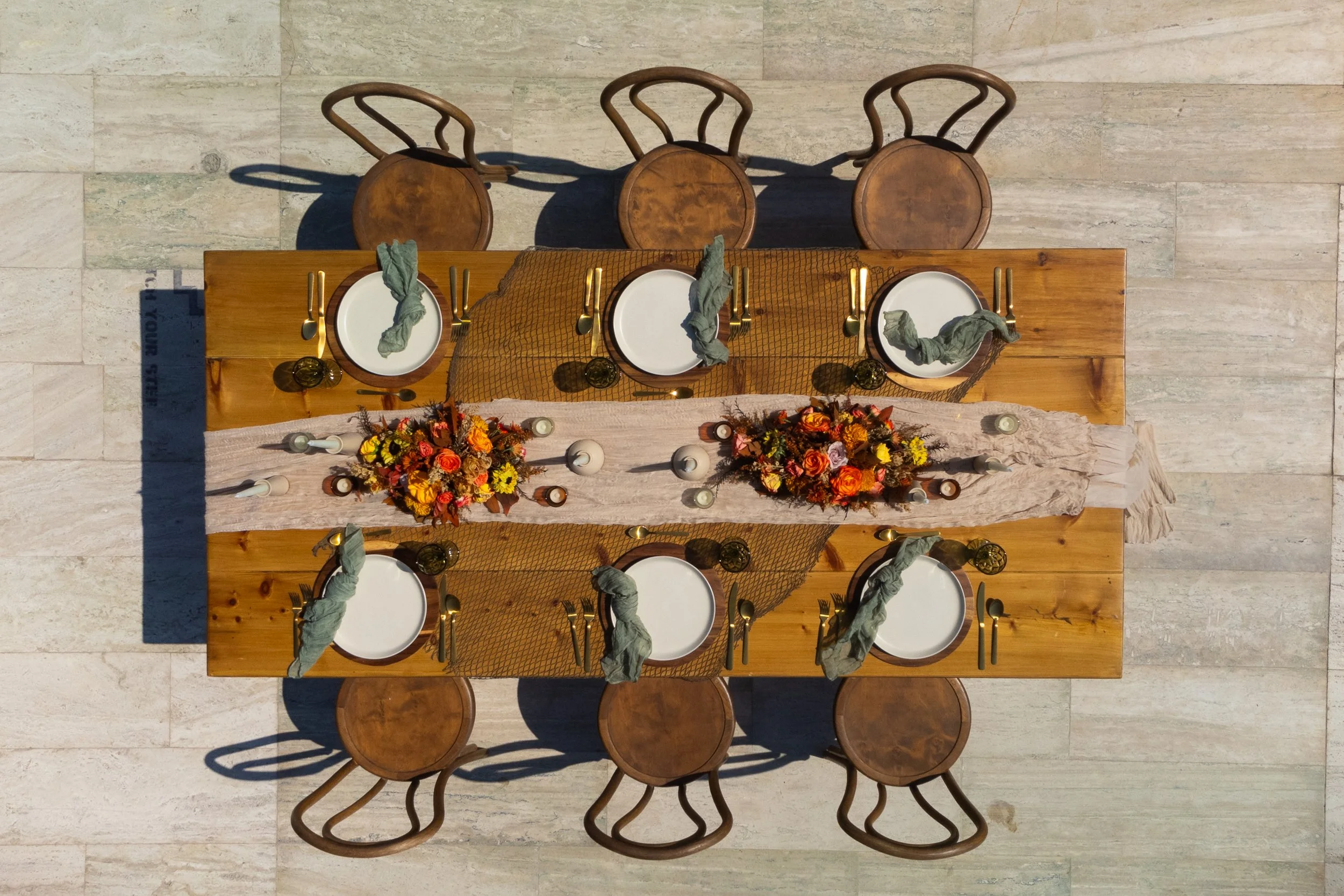 Top-down view of a wooden dining table set for six, decorated with floral arrangements and candles, with six matching wooden chairs around it, placed on a light-colored tile floor.