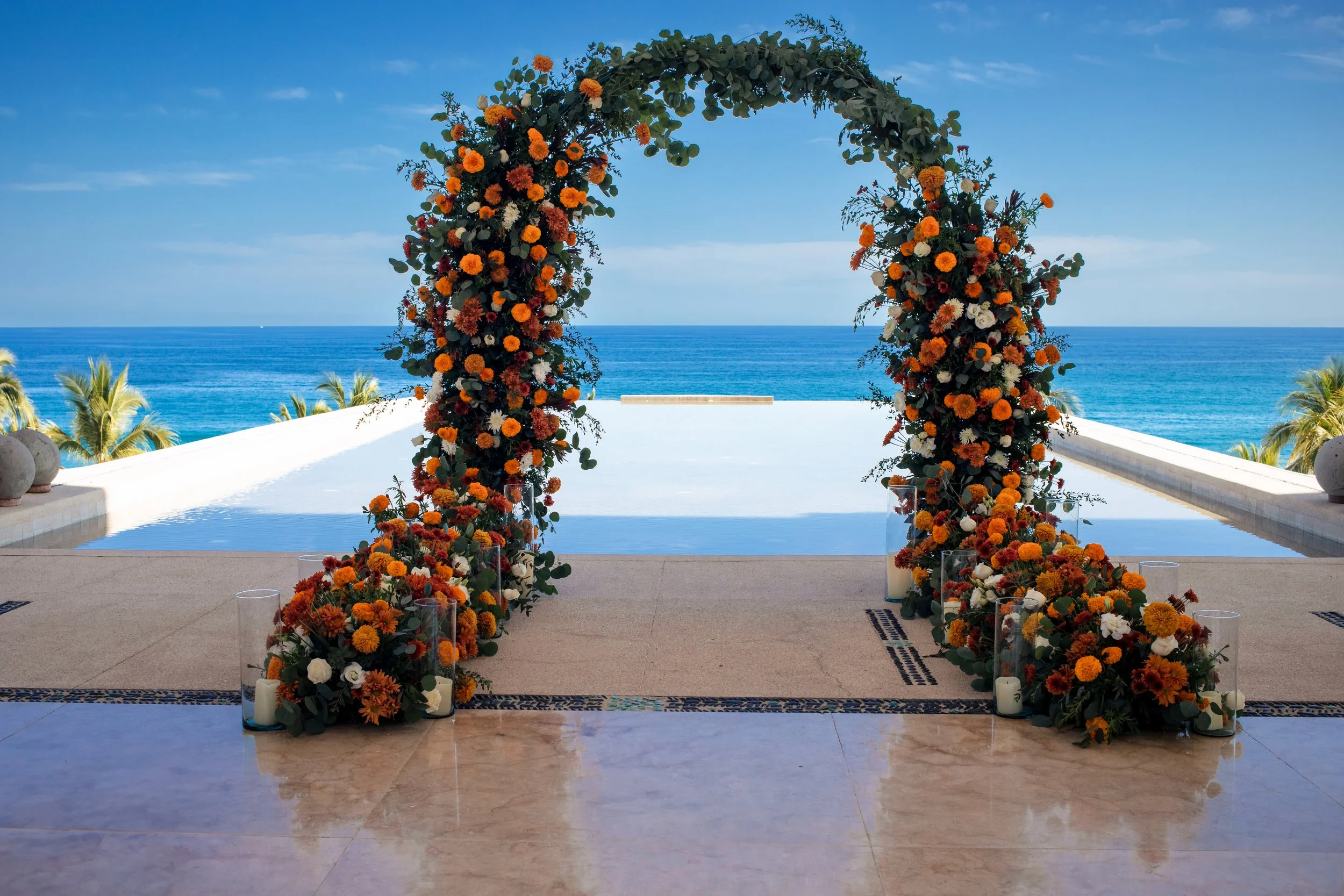 Oceanfront wedding ceremony arch in Los Cabos with orange, white, and red florals, greenery, and coastal wedding decor by I DO Weddings Los Cabos.