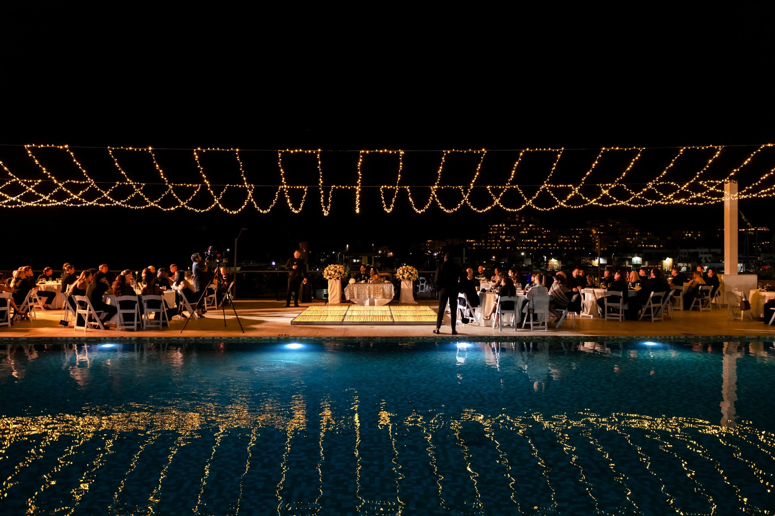 Nighttime outdoor event with round tables, white chairs, and floral centerpieces, decorated with string lights overhead, reflecting in a nearby pool, with people seated and a few standing, in a cityscape setting.