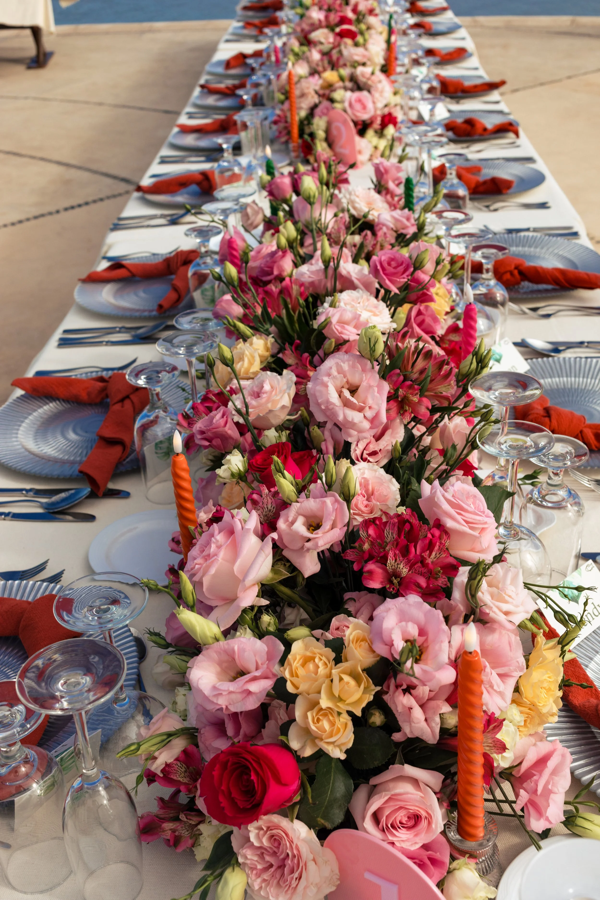 A long table decorated with a pink, peach, and red floral centerpiece, surrounded by place settings with white plates, silverware, glasses, and orange napkins, set outdoors near a body of water.