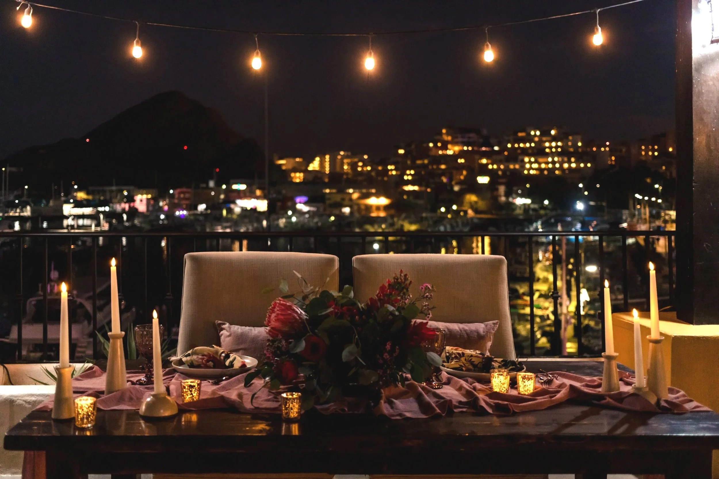 Outdoor dining table at night with candles, flowers, and a long tablecloth, overlooking a cityscape with illuminated buildings and a mountain in the background, string lights hanging overhead.