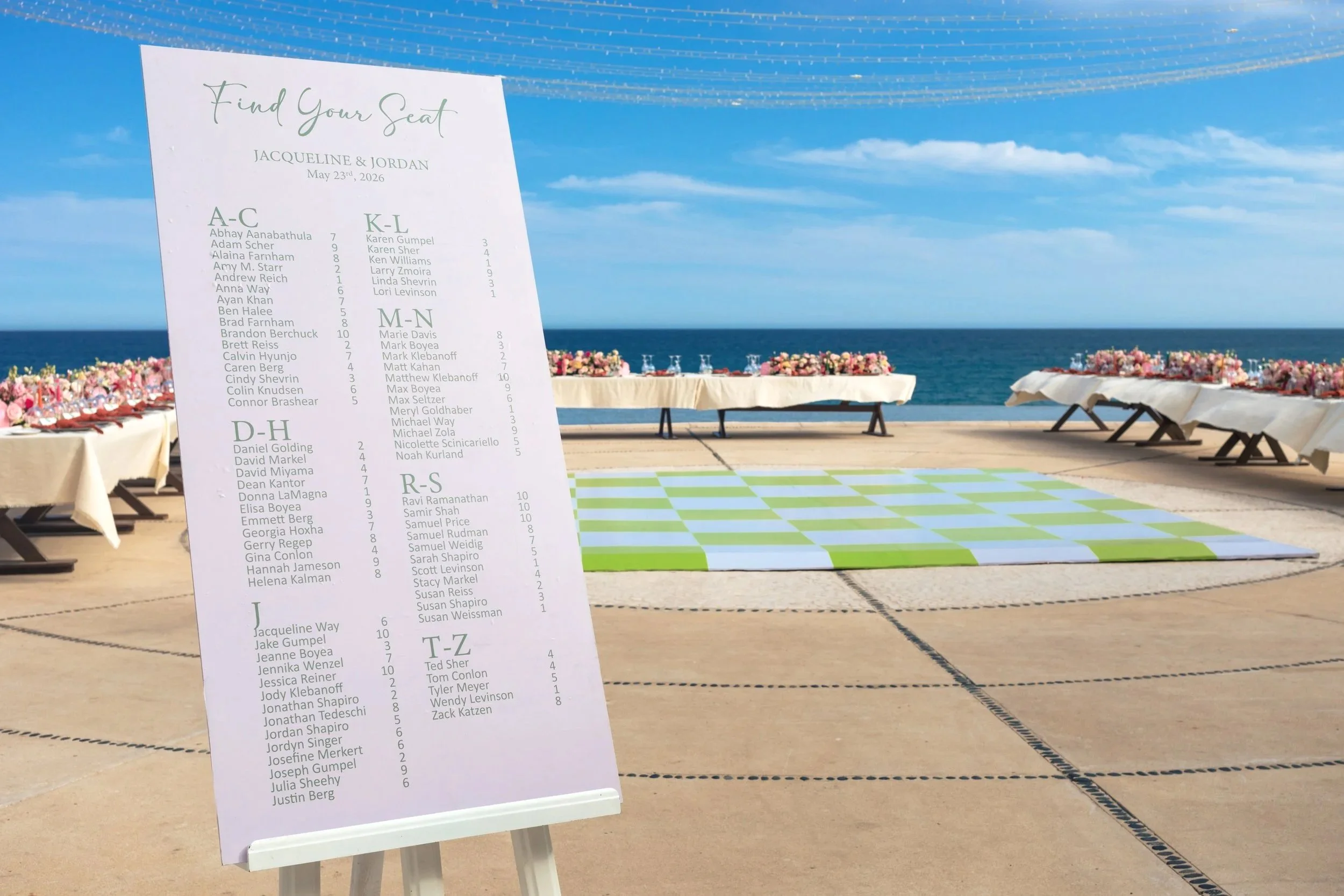 A wedding seating chart on a stand in front of a beach backdrop with tables decorated with pink flowers and covered in white tablecloths.