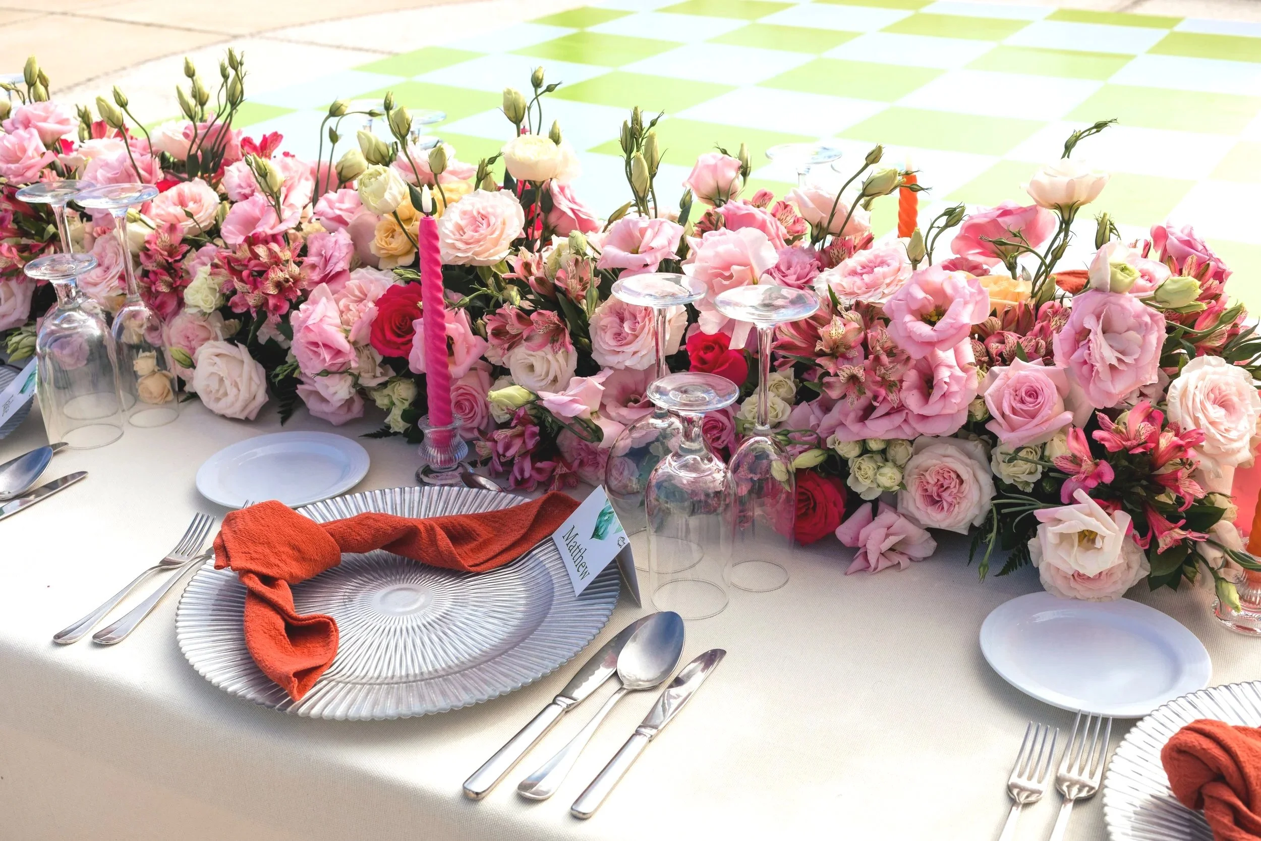 A table set for a special occasion with a large floral centerpiece of pink and white flowers, clear wine glasses, silverware, and orange cloth napkins.