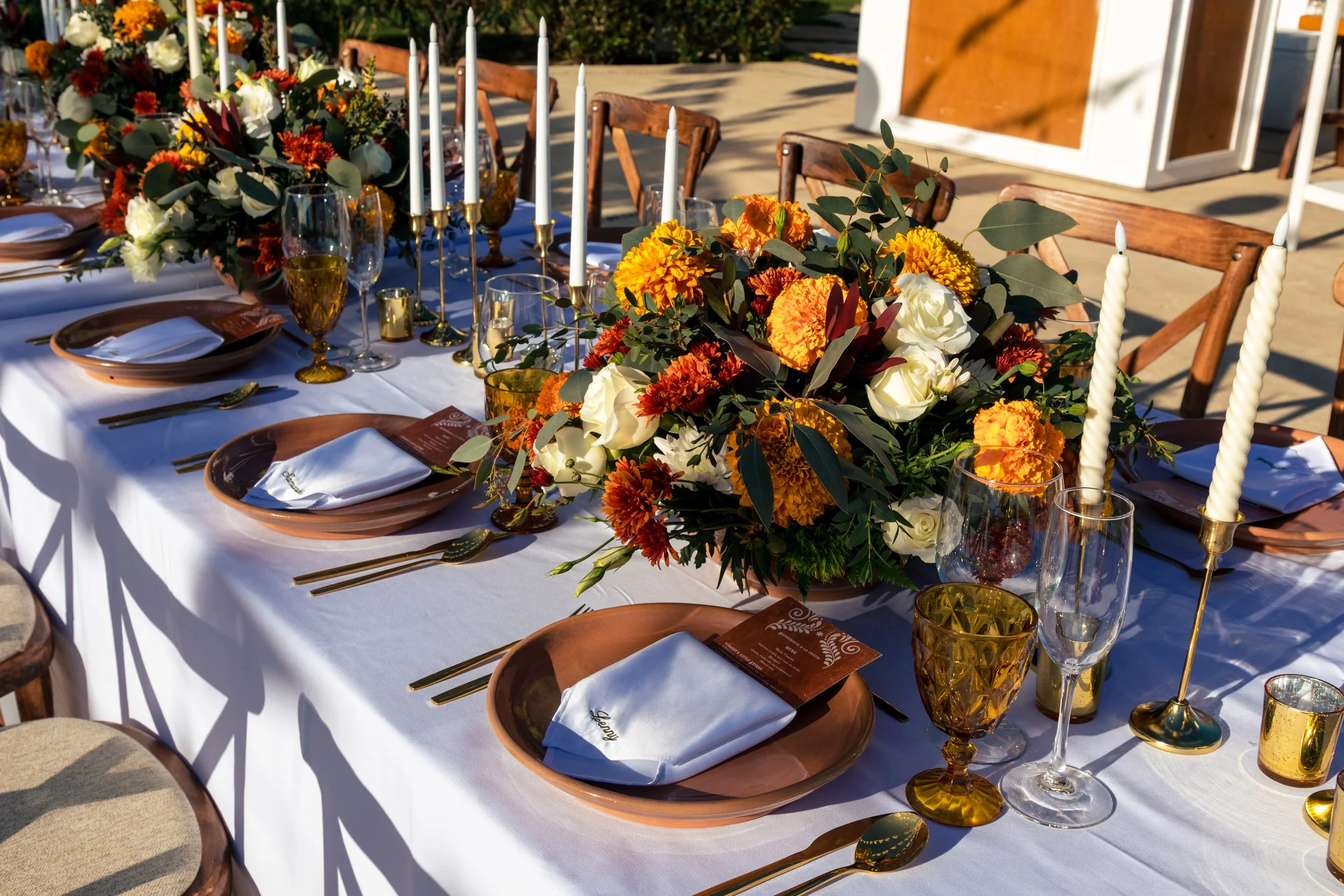A beautifully decorated outdoor wedding or event table with floral centerpieces in shades of orange, yellow, and white, set with gold-accented glassware, white candles, and elegant brown plates and napkins.