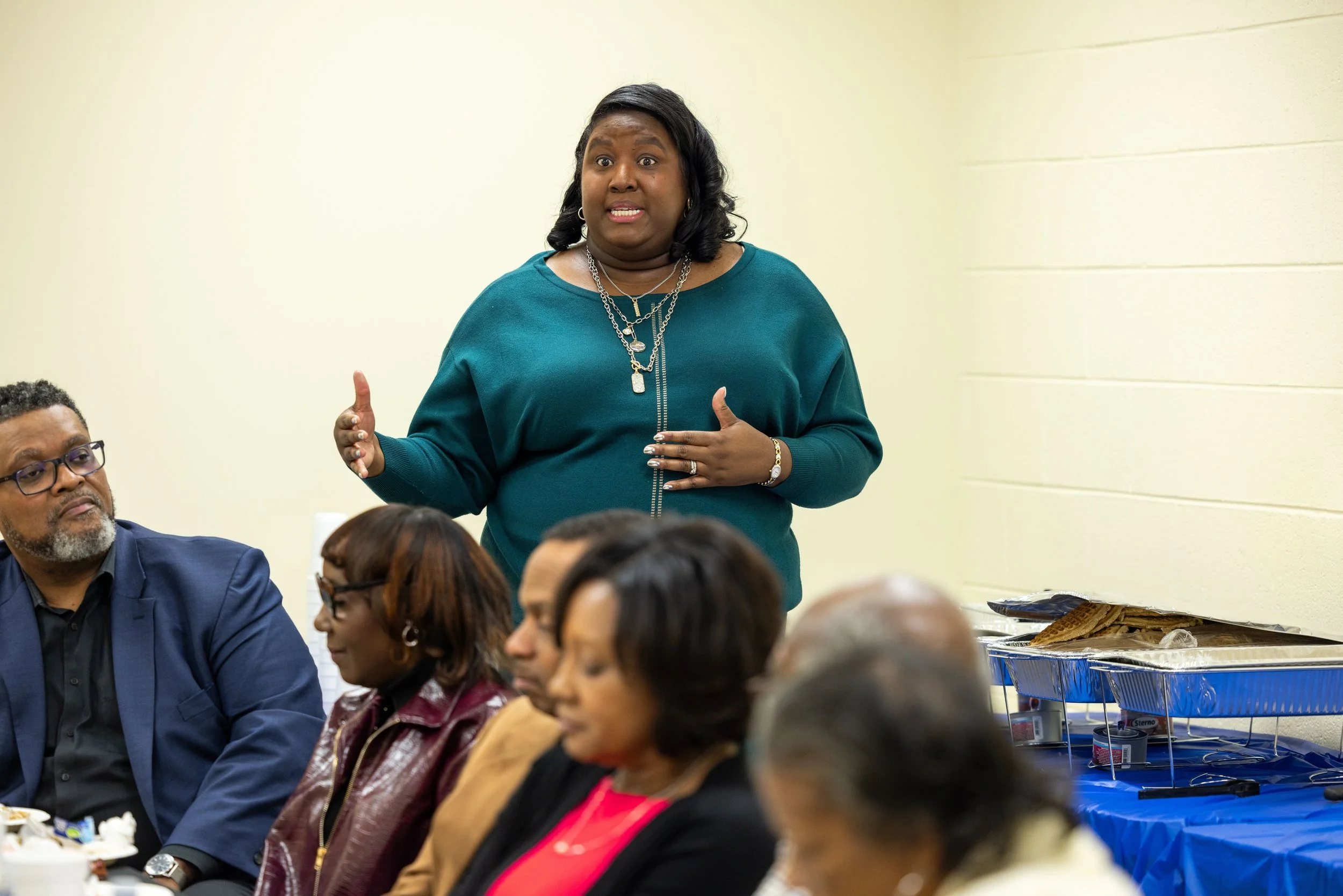 Monchiere' Holmes Jones, Cultural Economist™  standing and speaking during a community meeting, with several community leaders seated around a table, and trays of food in the background.