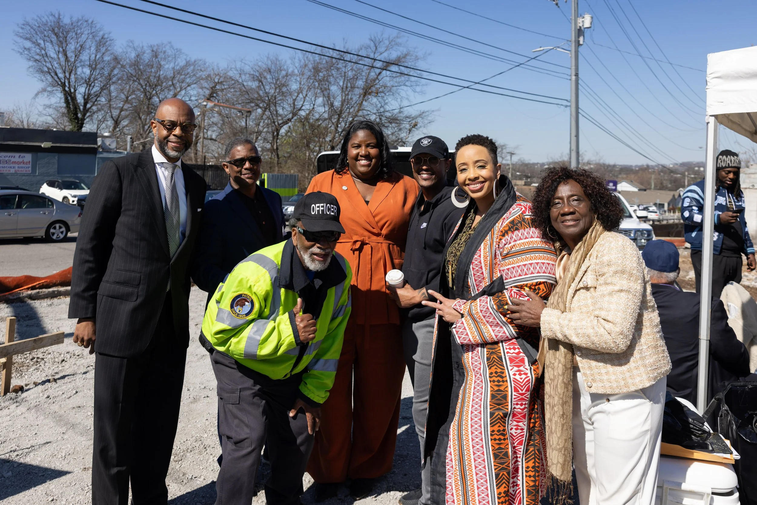 Monchiere' Holmes Jones, Cultural Economist™  with a group of community leaders smiling and posing outdoors during a community ground breaking event, some wearing jackets and sunglasses, with cars and trees in the background.