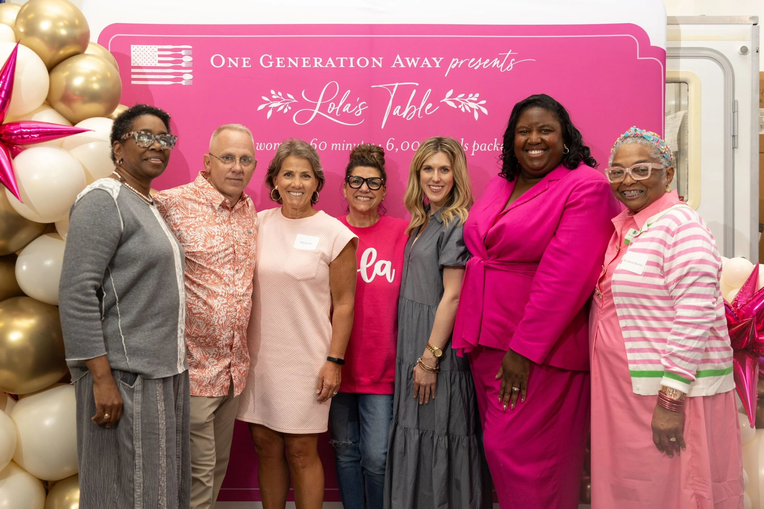 Monchiere' Holmes Jones, Cultural Economist™  with Group of women standing together at a community event with a pink backdrop and gold balloons. The backdrop reads "One Generation Away presents Lola's Table" and includes details about the event.