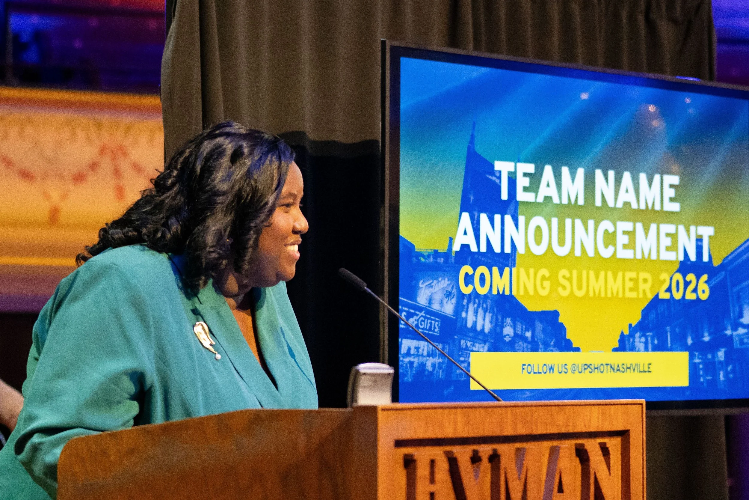Monchiere' Holmes Jones, Cultural Economist™  speaking at a press conference, at a podium with a large screen behind her displaying the text 'Team Name Announcement Coming Summer 2026' and a follow handle @UPSHOTNASHVILLE'.